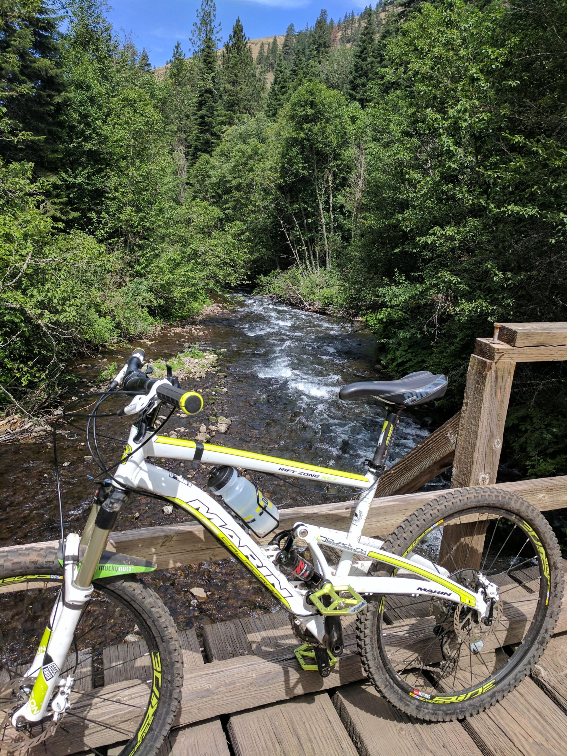 Marin Rift Zone: A white mountain bike with green and black accents is parked on a wooden bridge overlooking a flowing stream. The surroundings are lush with dense trees and greenery, and a clear blue sky is visible in the background.