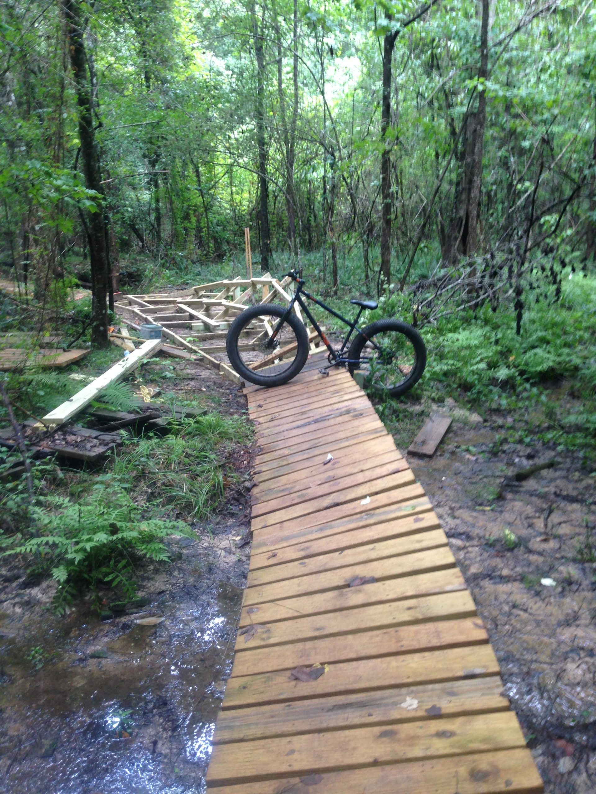 A wooden bike path winding through a lush, green forest, with a black fat tire bike leaning against the side. The path is elevated above a small stream, and construction materials can be seen in the background. Mt. Zion Bike Trails mountain bike trail.