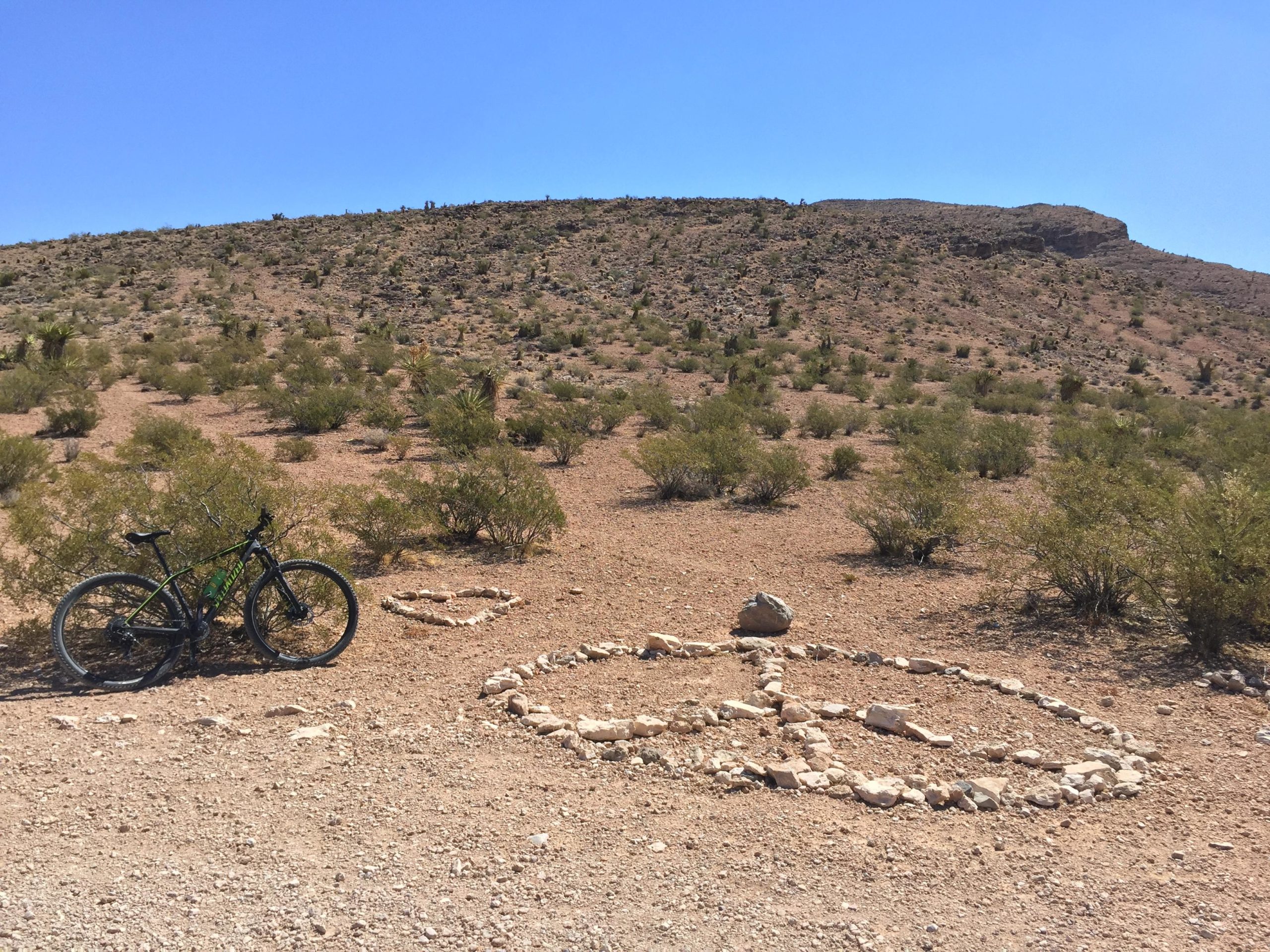 A mountain bike is leaning against a bush in a desert landscape, with a peace sign made of white stones on the ground nearby. The background features a rocky hillside under a clear blue sky. Sparse vegetation surrounds the area. Blue Diamond mountain bike trail.