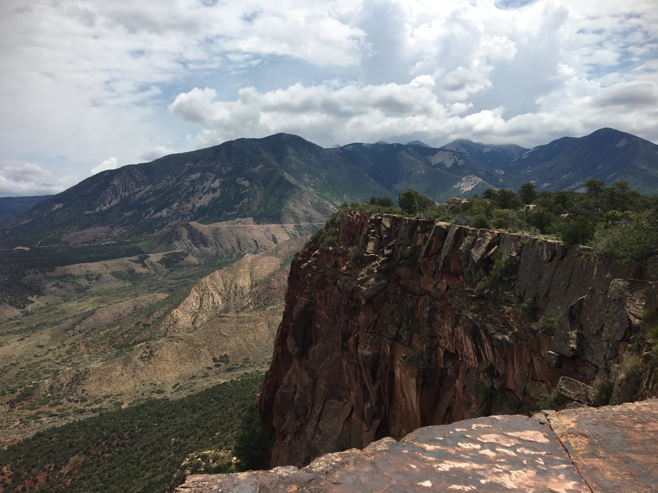 A panoramic view of rugged mountains and rolling hills under a partly cloudy sky. In the foreground, a rocky cliff edge provides a dramatic drop, with vibrant green vegetation scattered throughout the landscape. The background features a range of mountains with varying textures and colors, highlighting the natural beauty of the terrain. The Whole Enchilada mountain bike trail.