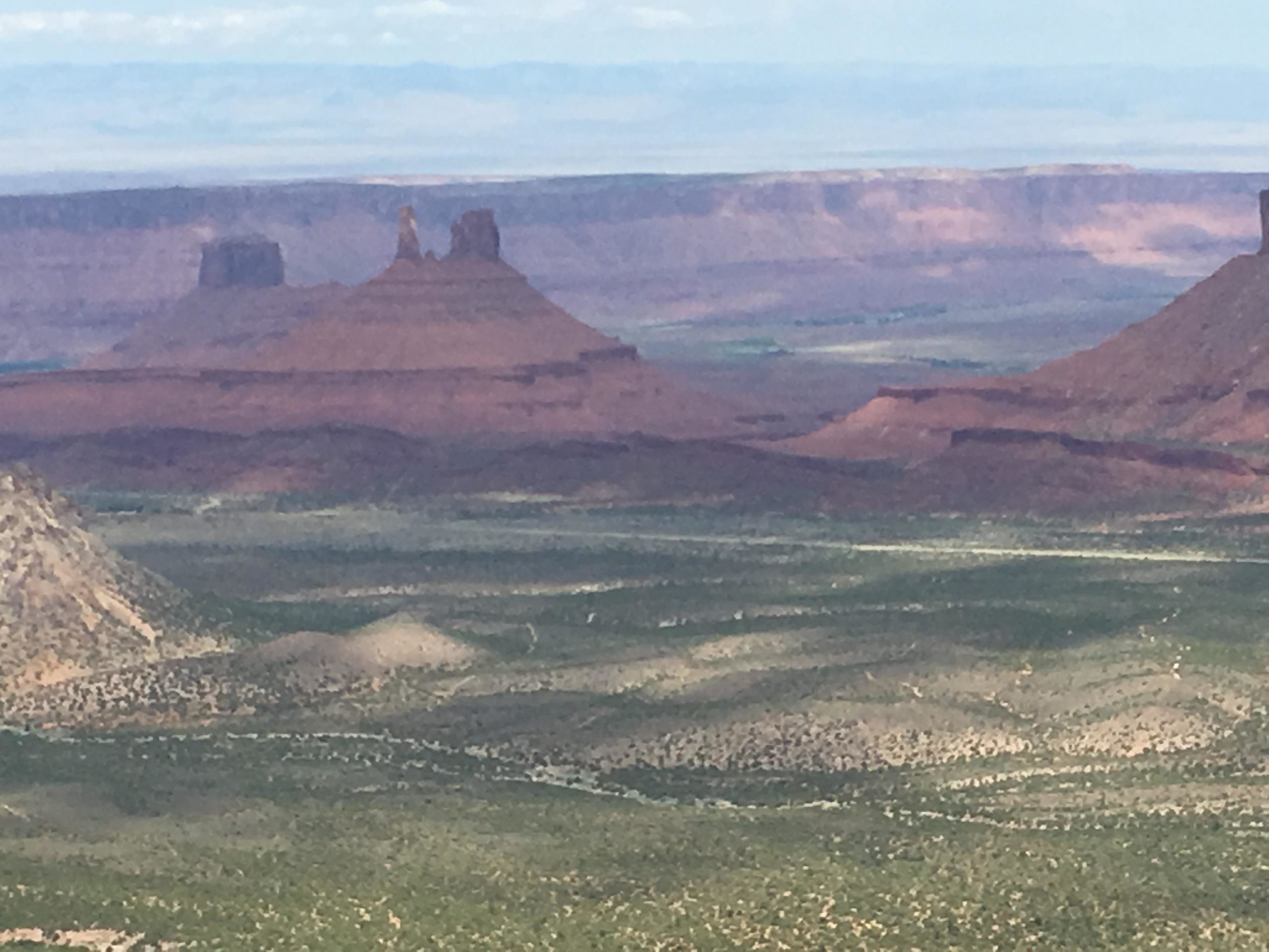 A panoramic view of a desert landscape featuring distinctive rock formations and mesas, with layers of red and brown rock under a blue sky. The foreground shows a mix of greenery and rugged terrain, while the distant mountains fade into the horizon. The Whole Enchilada mountain bike trail.