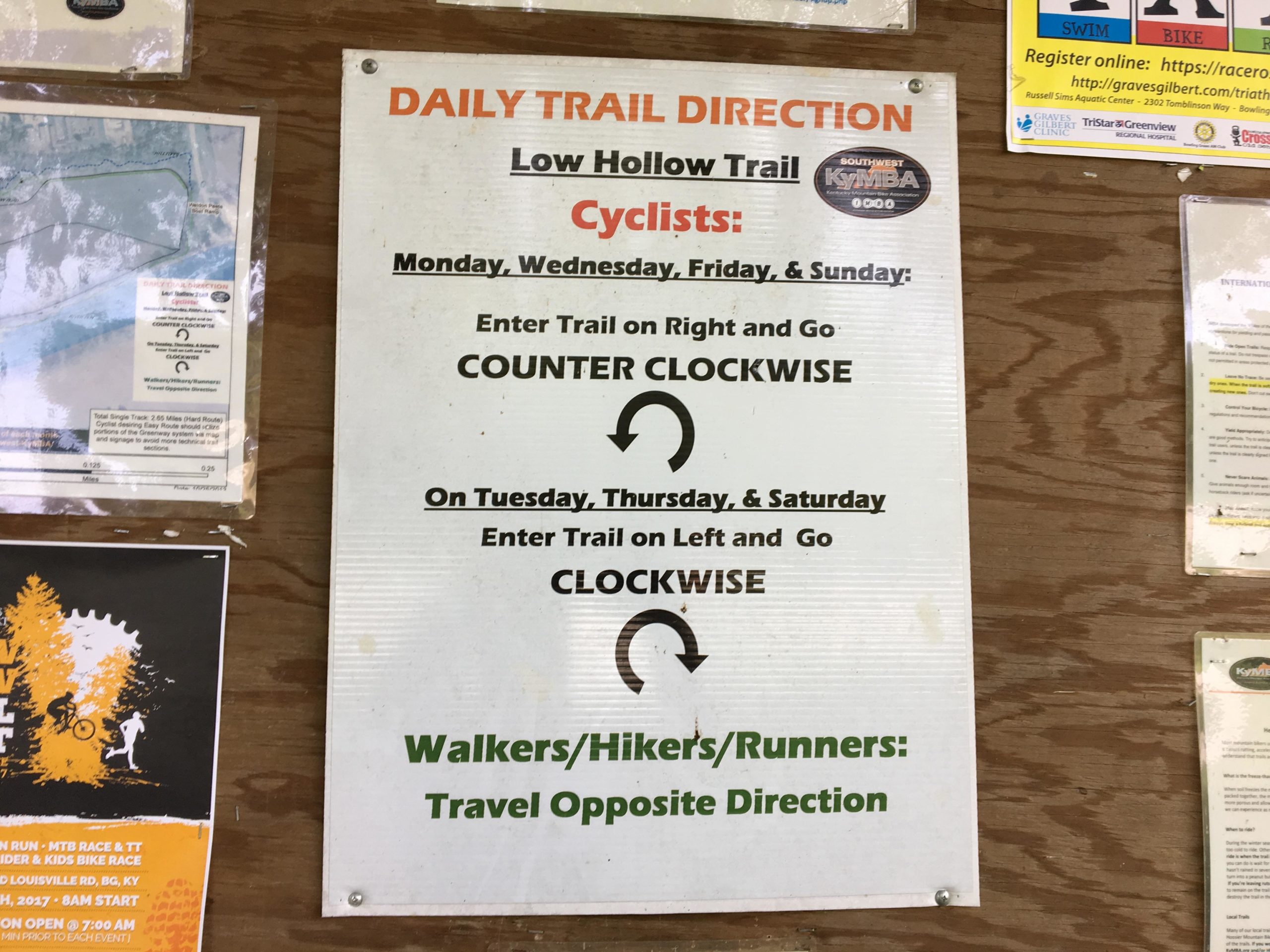Sign displaying trail directions for the Low Hollow Trail, including guidelines for cyclists and walkers/hikers/runners. It specifies the days cyclists should enter the trail and the direction to travel. The sign also features a map and other notices related to local events and activities. Low Hollow mountain bike trail.