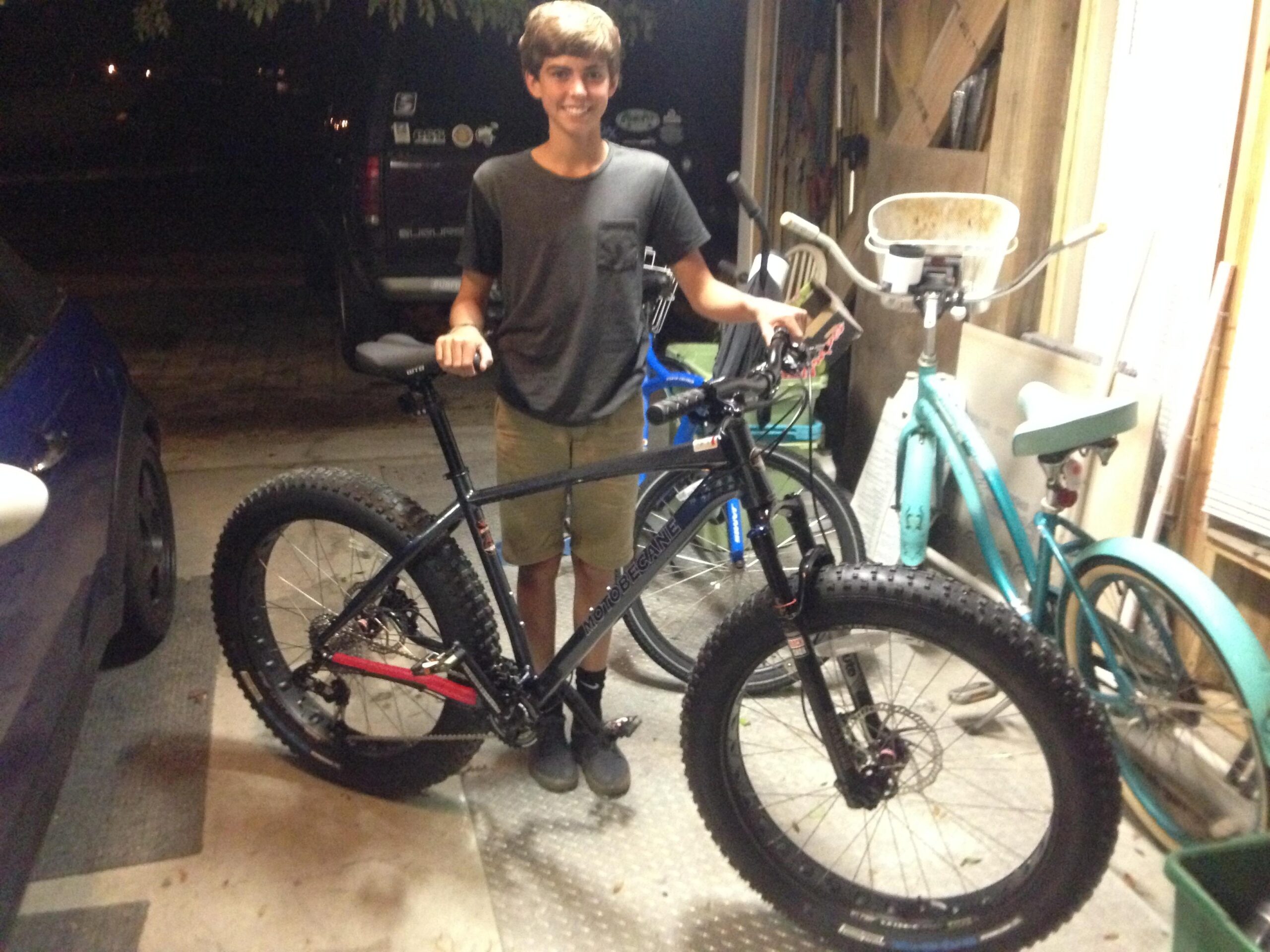 Motobecane Sturgis Bullet: A teenager stands in a dimly lit garage next to a large fat bike with wide tires. The boy is wearing a gray t-shirt and shorts, smiling as he holds onto the bike. Behind him, there are other bicycles and various garage items visible in the background.