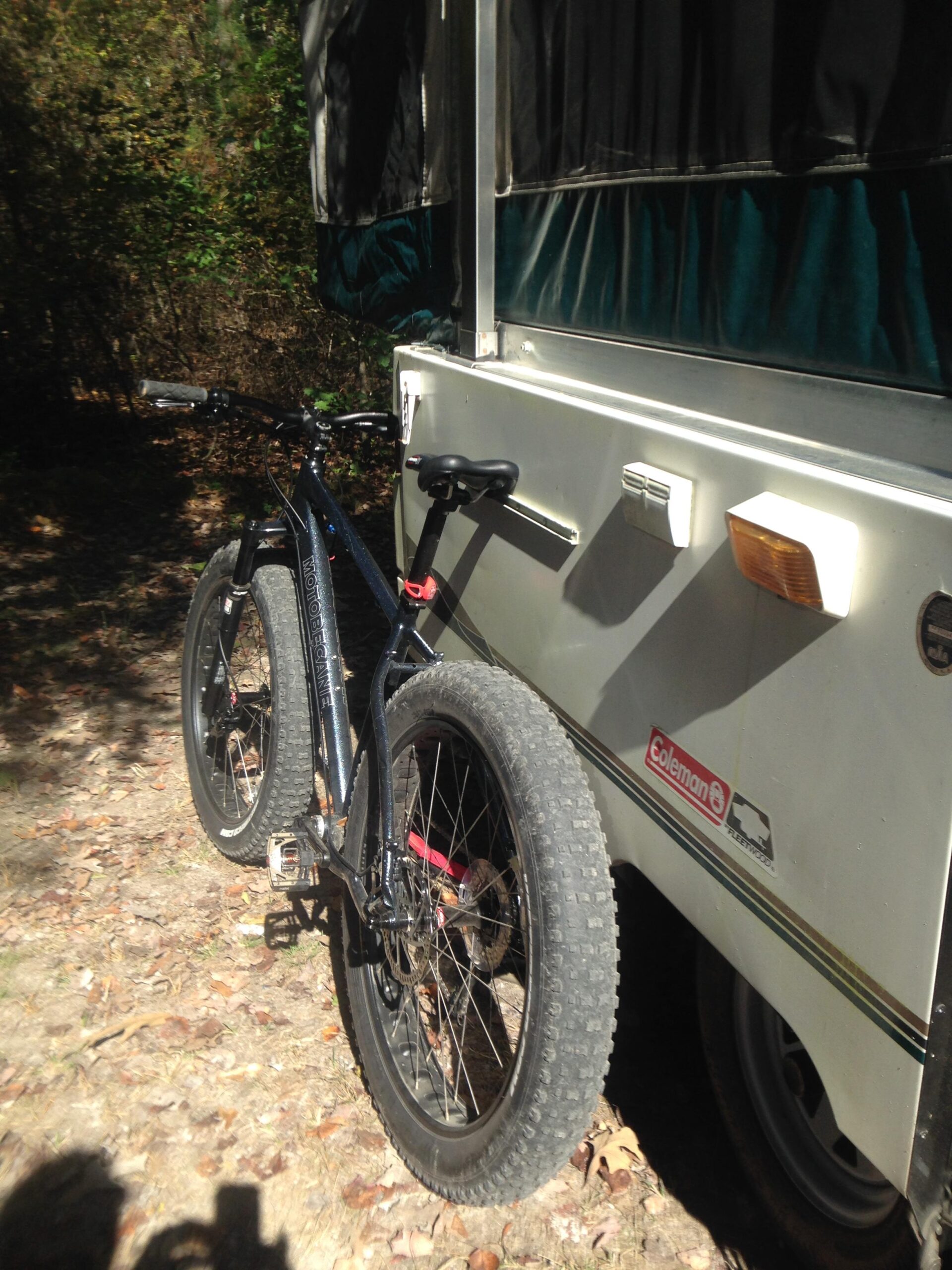 Motobecane Sturgis Bullet: A black mountain bike with fat tires is parked next to a white pop-up camper in a wooded area, surrounded by fallen leaves and greenery.