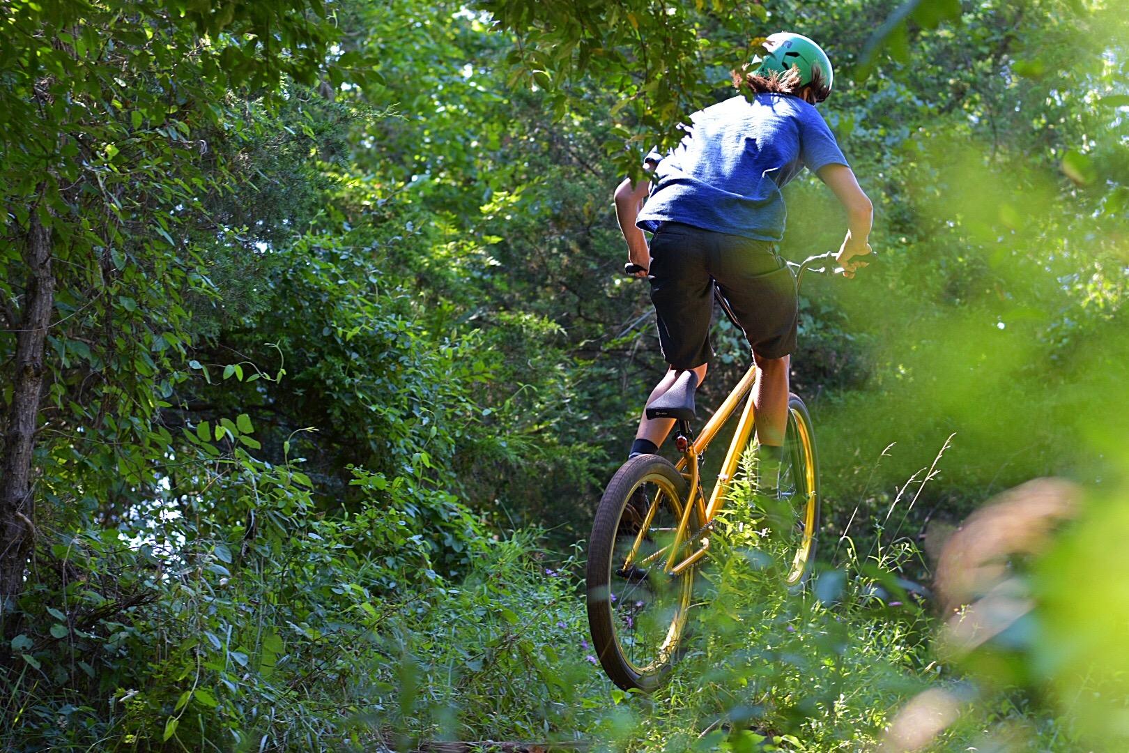 A person riding a yellow bicycle on a dirt path through a dense green forest. The rider is wearing a helmet and is slightly airborne as they navigate the terrain, surrounded by lush foliage. Six Mile Run mountain bike trail.