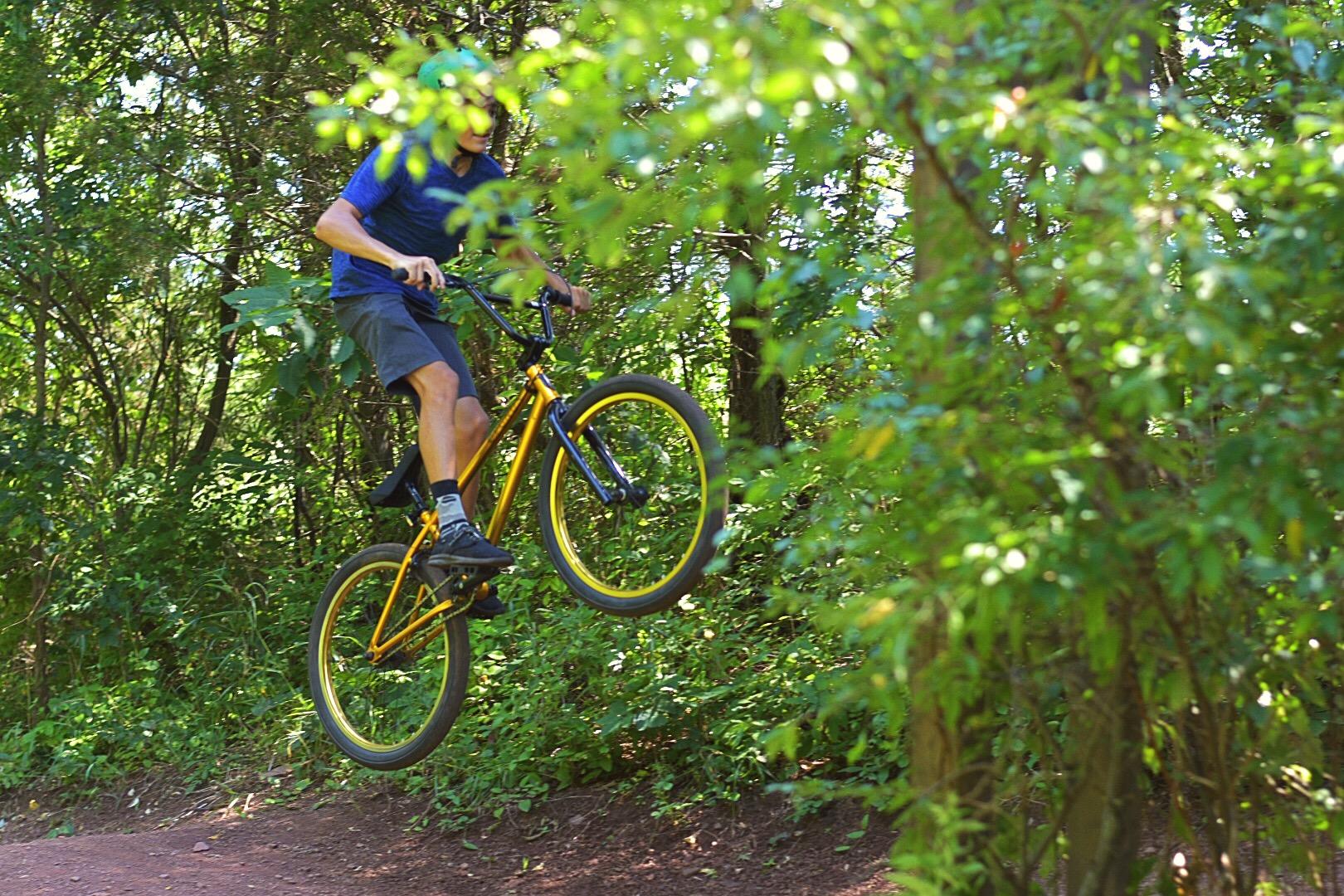 A cyclist in a blue shirt and helmet performs a jump on a yellow bike along a dirt trail surrounded by green foliage and trees. Six Mile Run mountain bike trail.
