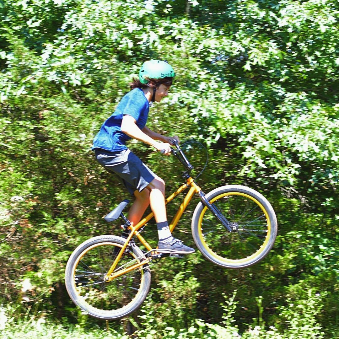 A young boy in a green helmet rides an orange bicycle, performing a jump on a dirt path surrounded by greenery. Six Mile Run mountain bike trail.