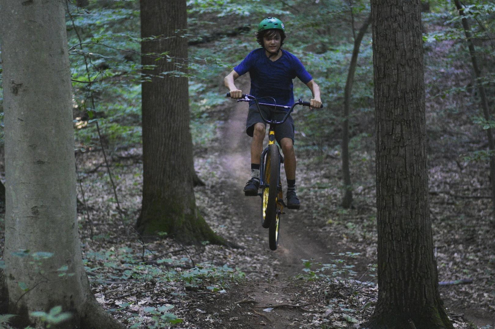 A young boy wearing a green helmet jumps over a dirt trail on his bike in a wooded area. Trees surround the path, and the scene is filled with green foliage and dappled sunlight. Clayton Park mountain bike trail.