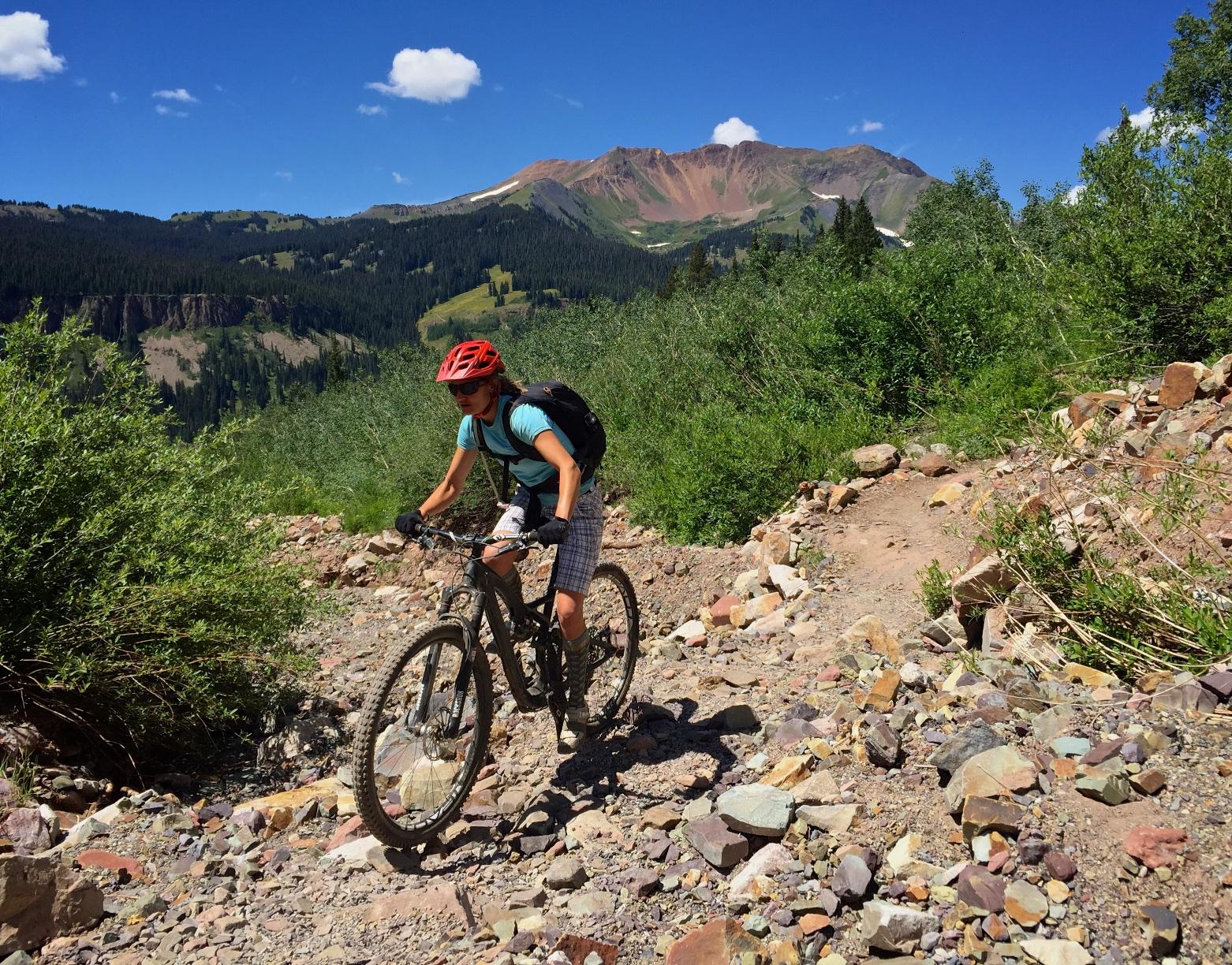 A mountain biker navigating a rocky trail surrounded by lush greenery and mountains under a clear blue sky. The cyclist is wearing a red helmet and has a backpack, focusing on the rough terrain ahead. Trail 401 mountain bike trail.