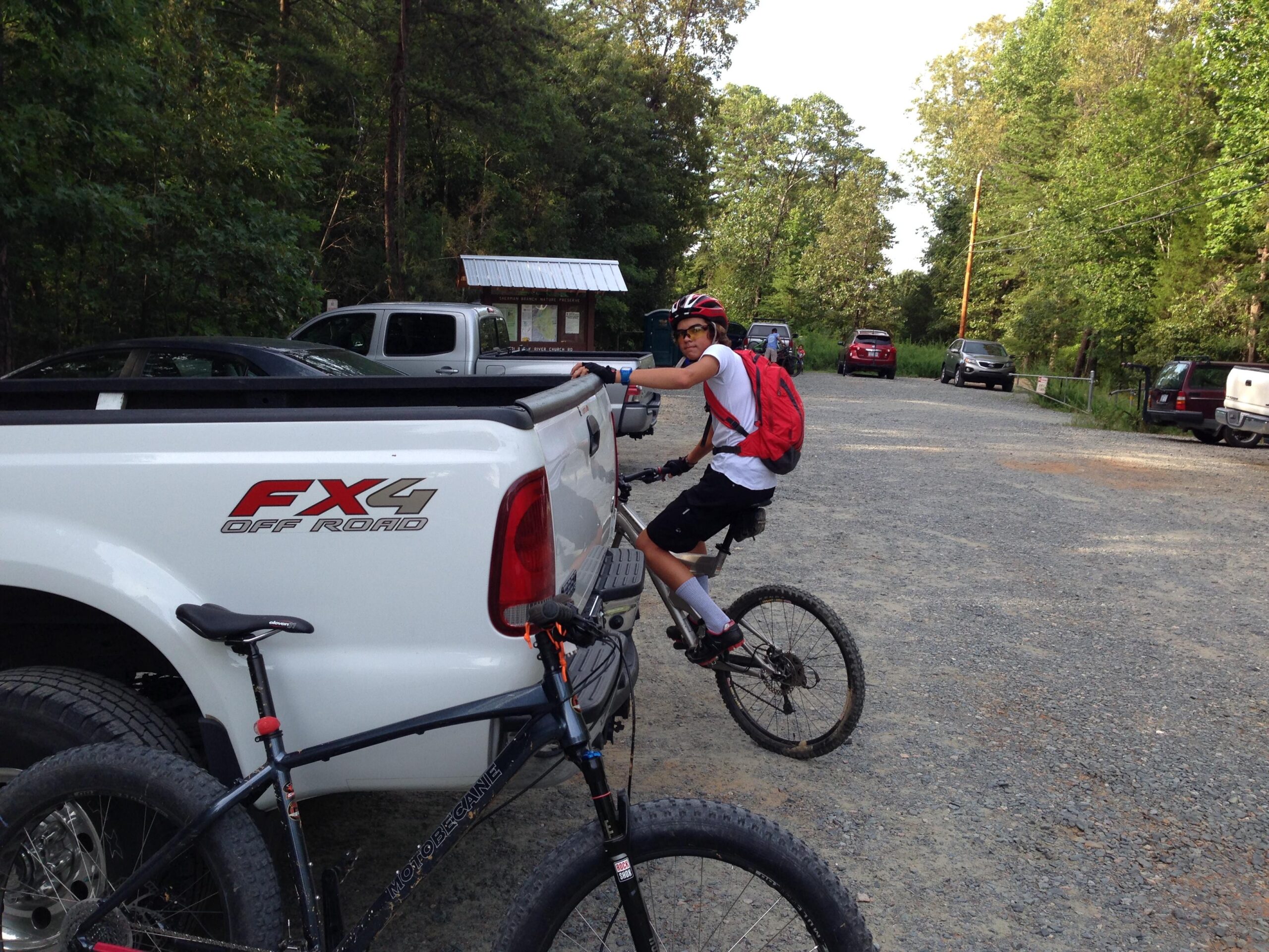 Motobecane Sturgis Bullet: A young person in a white t-shirt, black shorts, and a red backpack is sitting on a mountain bike and looking back towards the camera. In the background, a white off-road pickup truck and several other parked vehicles are visible, along with trees and a gravel parking area.