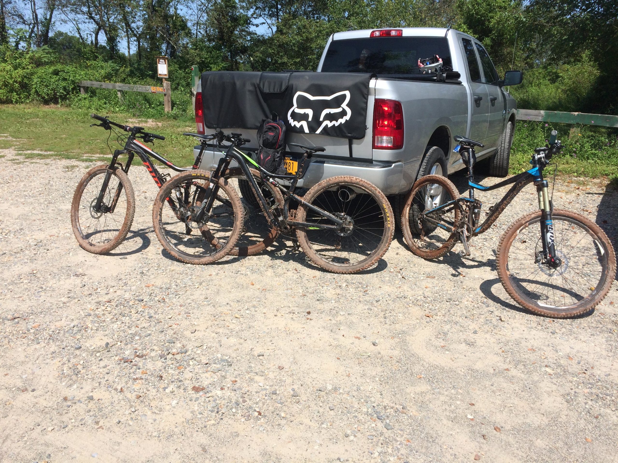 Alt text: "Four mountain bikes with muddy tires parked beside a silver pickup truck in a gravel area surrounded by greenery." Six Mile Run mountain bike trail.
