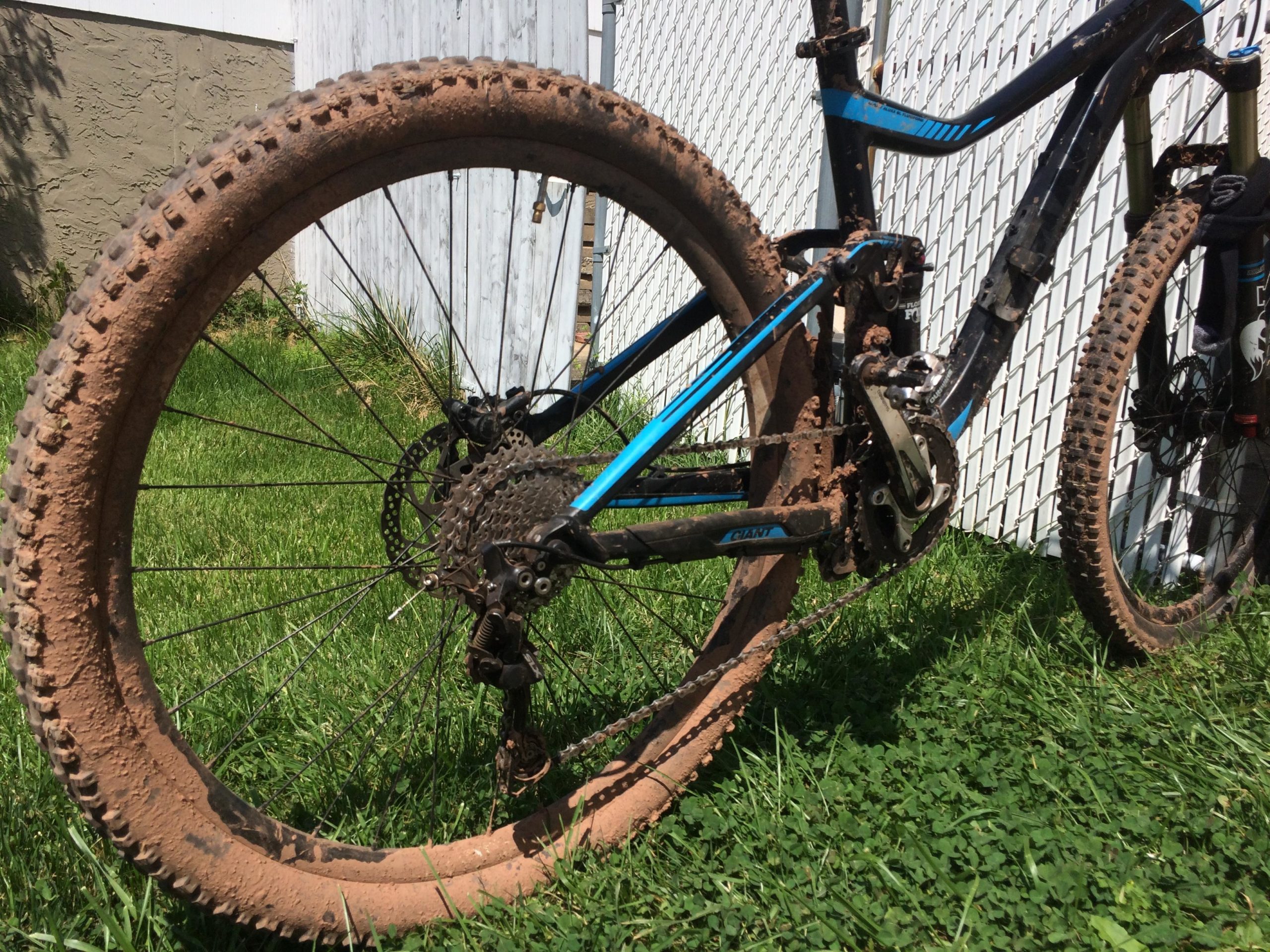A close-up view of a muddy mountain bike resting on green grass, showcasing its rear wheel and part of the frame. The bike is heavily coated in dirt, particularly on the tires and chain, indicating recent use on a muddy trail. A white fence and a textured wall are visible in the background. Six Mile Run mountain bike trail.