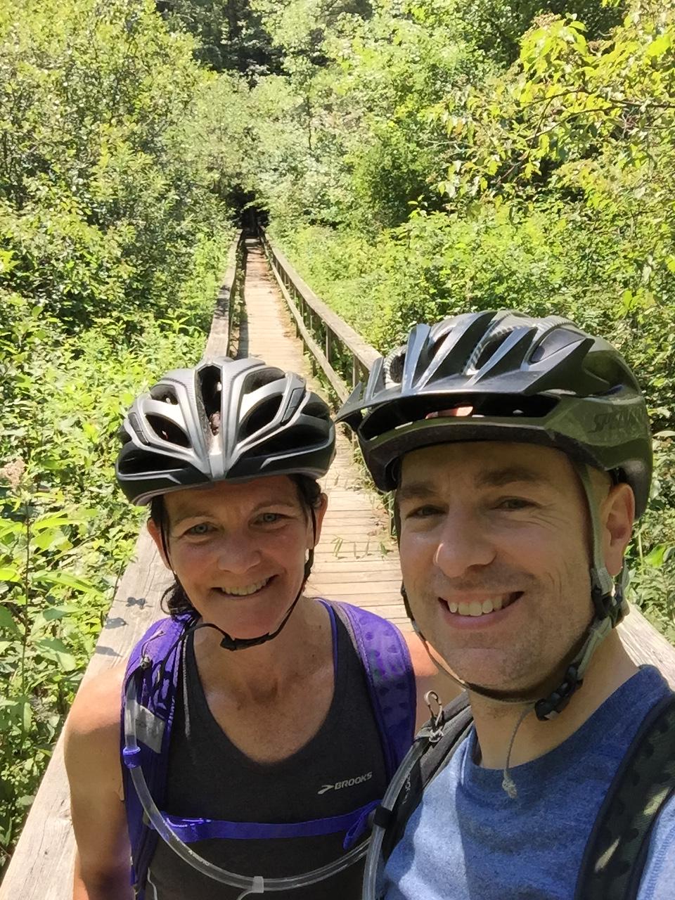 A man and a woman pose for a selfie while wearing cycling helmets, standing on a wooden boardwalk surrounded by lush greenery. The scene captures a sunny day in a natural setting, with trees and plants lining the path. Stonewall Farm Race Course mountain bike trail.