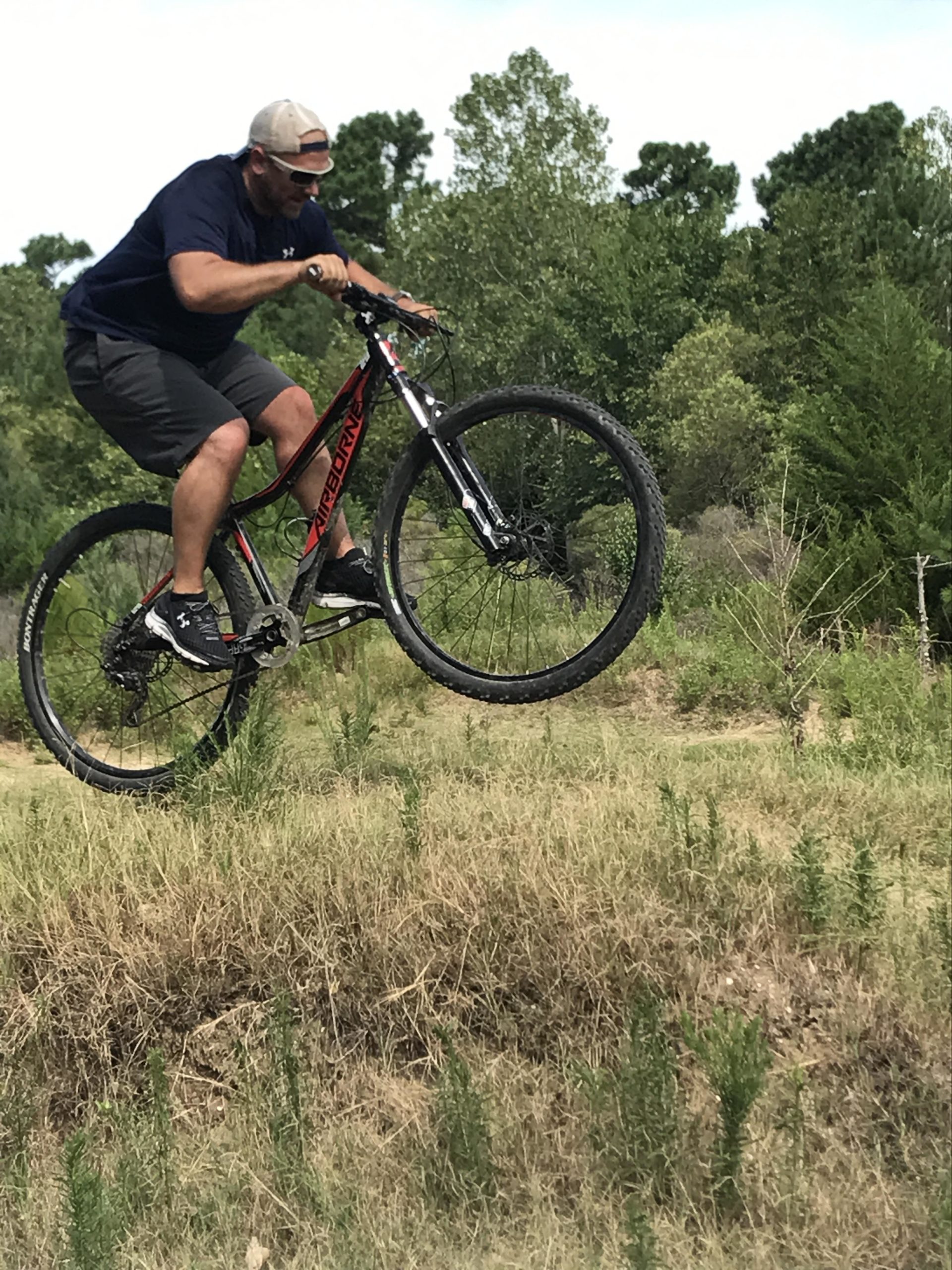 A man riding a mountain bike performs a jump over a small hill, surrounded by greenery and trees in the background. He wears a cap and sunglasses, and is focused on maintaining balance while in mid-air. Horry County Bike Run Park mountain bike trail.