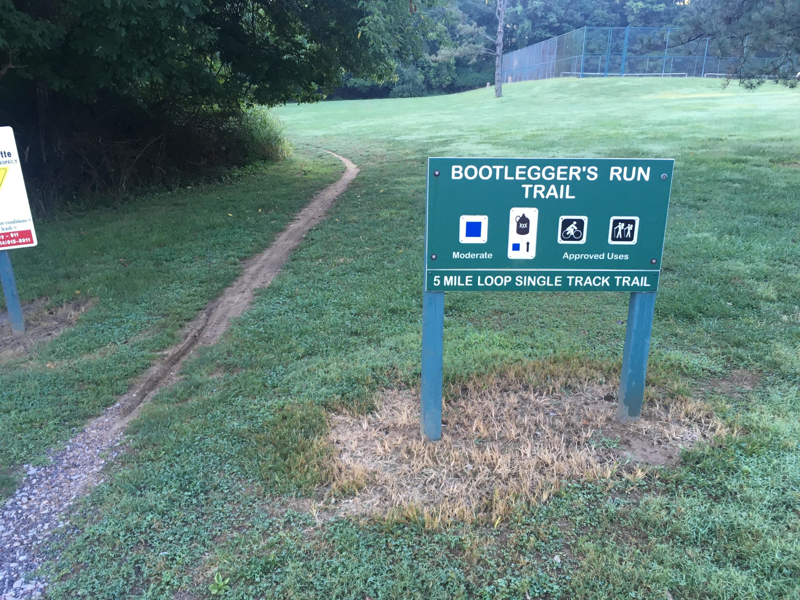 Sign marking the entrance to Bootlegger's Run Trail, featuring information about the trail's difficulty level (moderate), approved uses, and the length of the trail (5-mile loop single track). A narrow dirt path leads from the sign into a grassy area surrounded by trees. Creve Couer Park mountain bike trail.