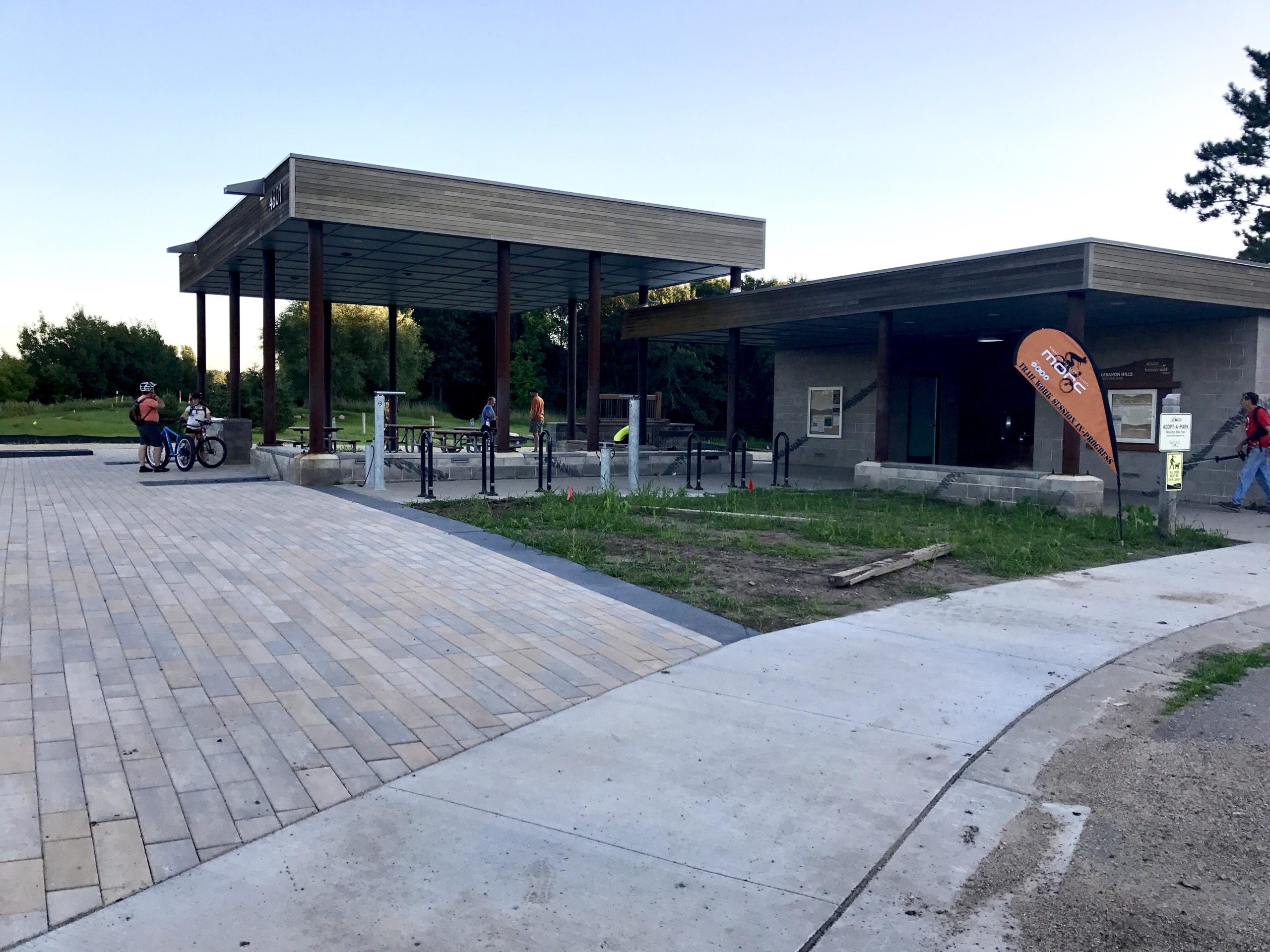 A modern outdoor facility featuring a wooden canopy and a stone building, with bike racks in the foreground. Several people are milling around, including two individuals on bicycles. An orange flag with a logo is visible, and the area is surrounded by greenery and pathways. The scene is set in the late evening with a clear sky. Lebanon Hills mountain bike trail.