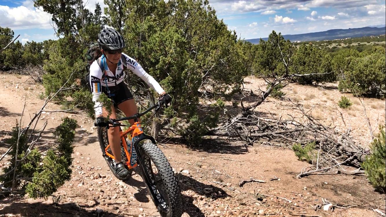 A person riding a fat bike on a rocky trail surrounded by shrubs and trees, with a clear blue sky and distant hills in the background. The rider is wearing a helmet and a cycling jersey, showcasing an action shot as they navigate the terrain. La Tierra mountain bike trail.