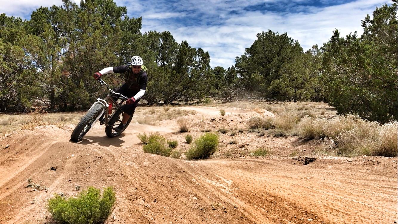 A cyclist in a helmet and gear leans into a turn while riding a fat bike on a dirt trail, surrounded by shrubs and trees under a partly cloudy sky. Dust is kicked up from the trail, emphasizing the motion and activity. La Tierra mountain bike trail.
