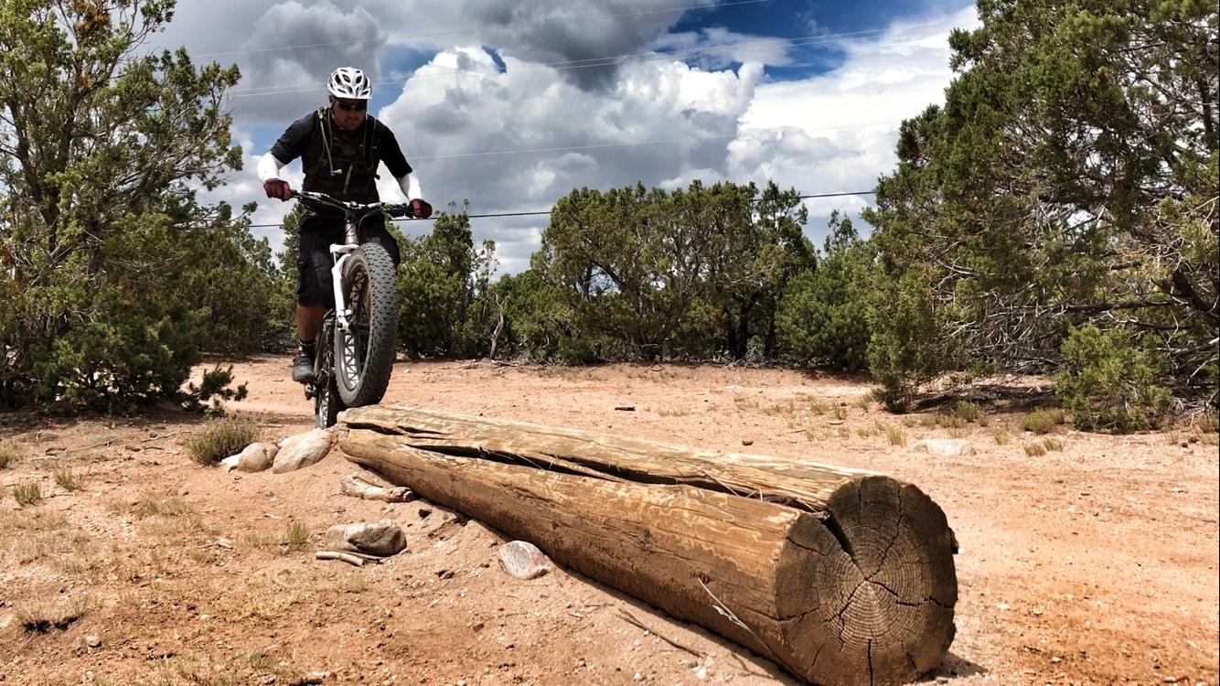 A person riding a fat bike performs a wheelie over a fallen log on a dirt trail surrounded by shrubs and trees under a partly cloudy sky. La Tierra mountain bike trail.
