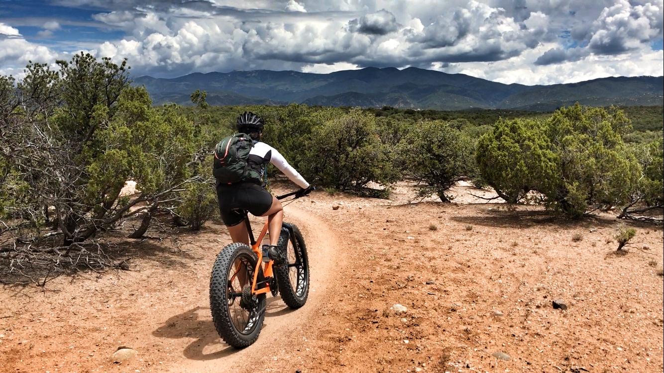 A person riding a fat tire bike on a sandy trail through a desert landscape, surrounded by shrubs and distant mountains under a partly cloudy sky. La Tierra mountain bike trail.