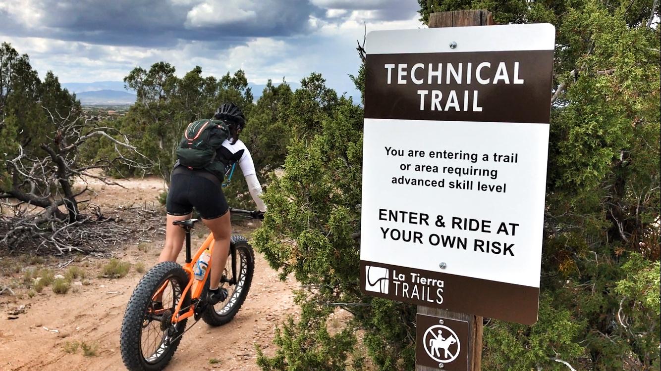 A mountain biker approaches a sign that reads "Technical Trail," indicating that the trail requires advanced skill level. The sign advises riders to enter and ride at their own risk. The trail is surrounded by trees, and the sky is partly cloudy. La Tierra mountain bike trail.