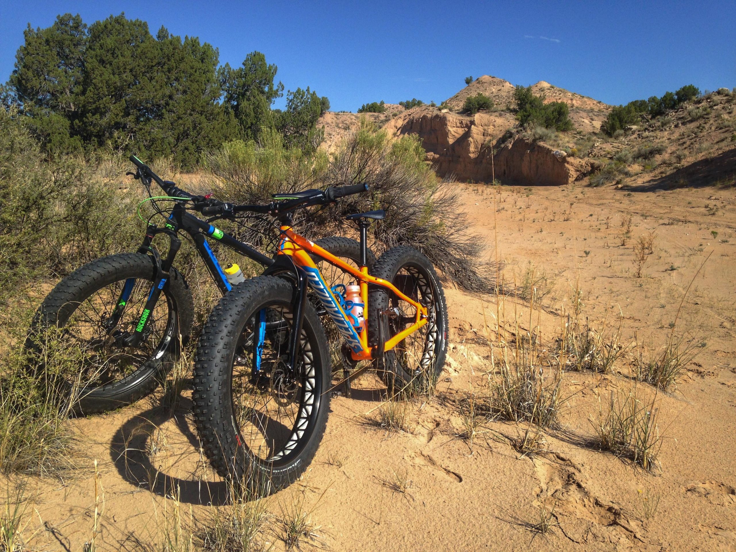 Two mountain bikes, one blue and black and the other orange, are leaning against a bush in a sandy outdoor landscape. The background features a rocky terrain with scattered vegetation under a clear blue sky. Mariposa Fat Bike Trails mountain bike trail.
