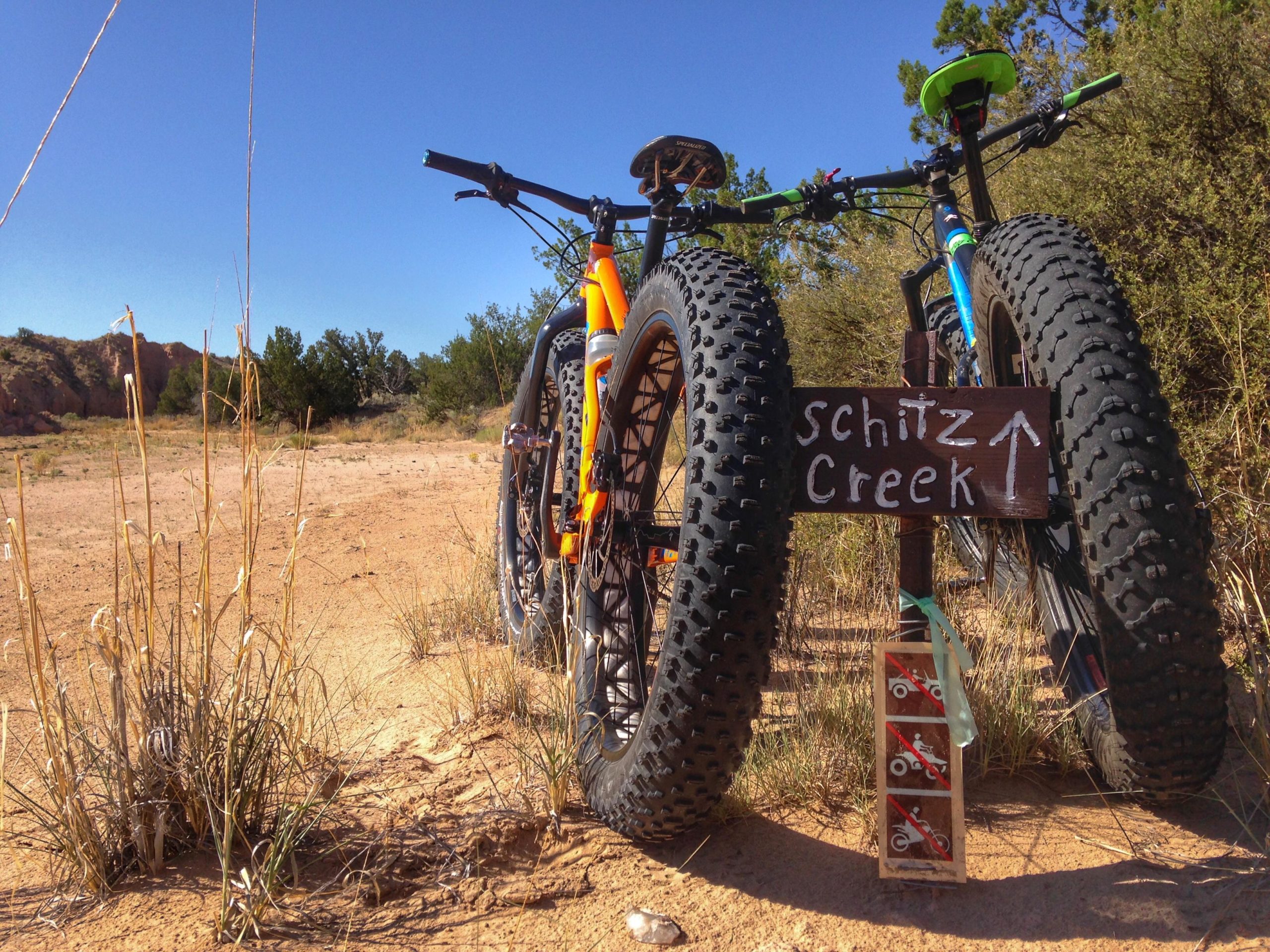 Two fat-tire bicycles lean against a wooden sign directing to "Schitz Creek," set against a backdrop of sandy terrain and sparse vegetation under a clear blue sky. The sign features symbols indicating restrictions on motor vehicles and cyclists. Mariposa Fat Bike Trails mountain bike trail.