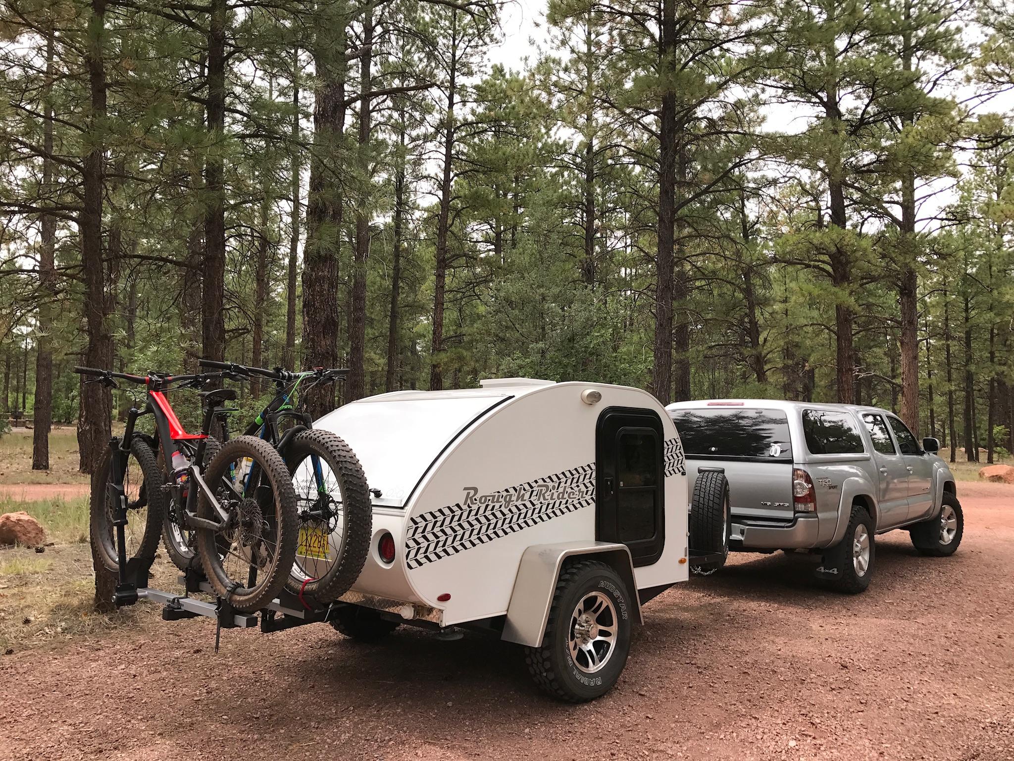 A compact white trailer with a tire tread design hitched to a silver pickup truck, parked in a forested area. Two mountain bikes are secured on a rack attached to the trailer. The background features tall pine trees and a dirt road, creating a peaceful outdoor setting. Hilso Mountain Bike Trail mountain bike trail.