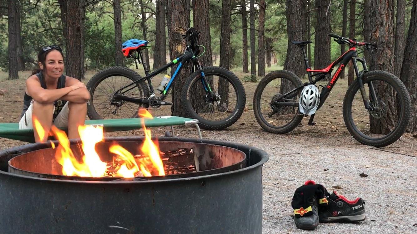 A person sitting by a campfire in a forested area, with two mountain bikes visible in the background. The individual is wearing casual clothing and appears relaxed, while flames from the fire flicker in the foreground. Nearby, a pair of cycling shoes is placed on the ground. Hilso Mountain Bike Trail mountain bike trail.