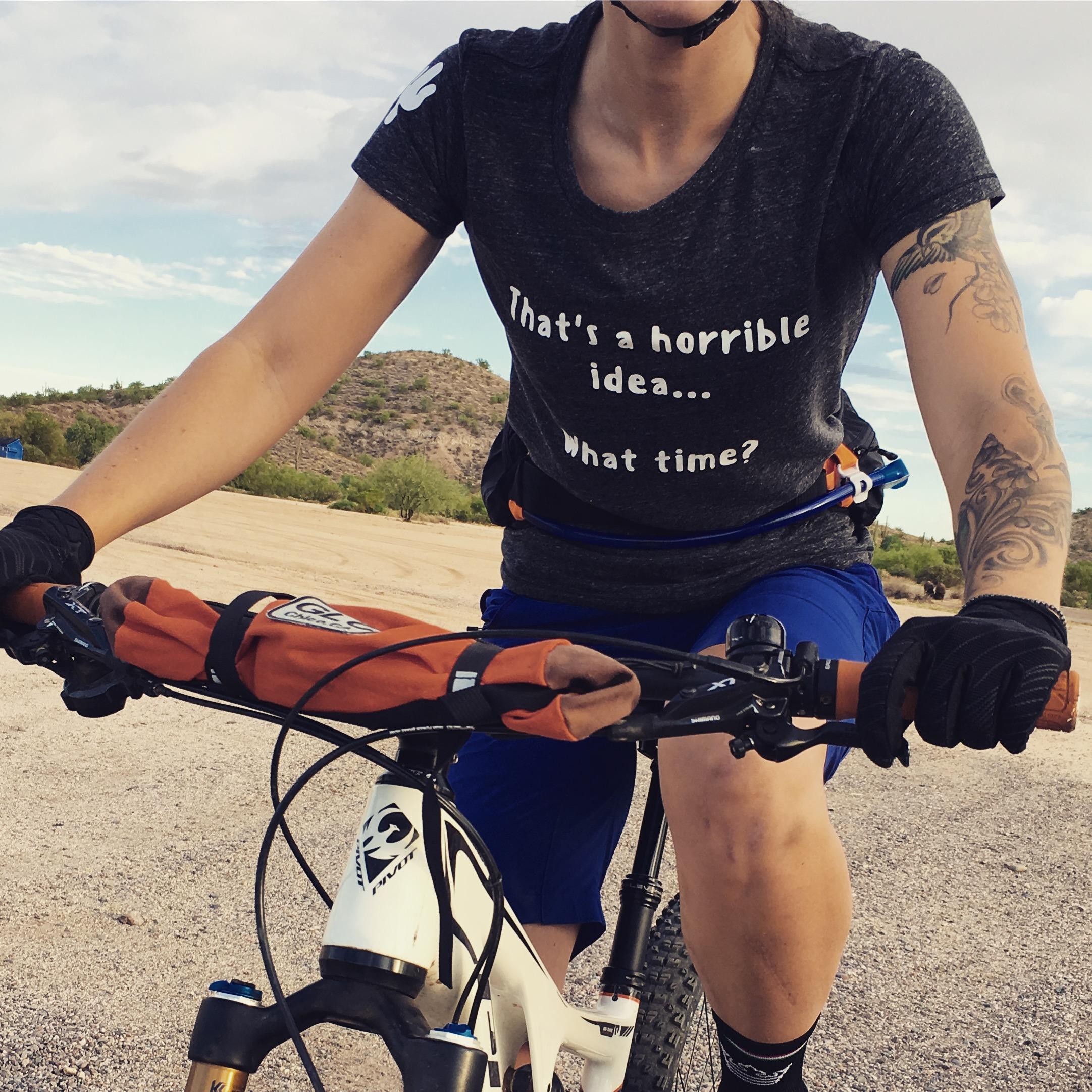 A person wearing a black t-shirt with white text reading "That's a horrible idea... What time?" is sitting on a mountain bike. They have short blue shorts, gloves, and visible tattoos on their arm. The background features a sandy area with a hill and sparse vegetation under a partly cloudy sky. McDowell Mountain Park mountain bike trail.