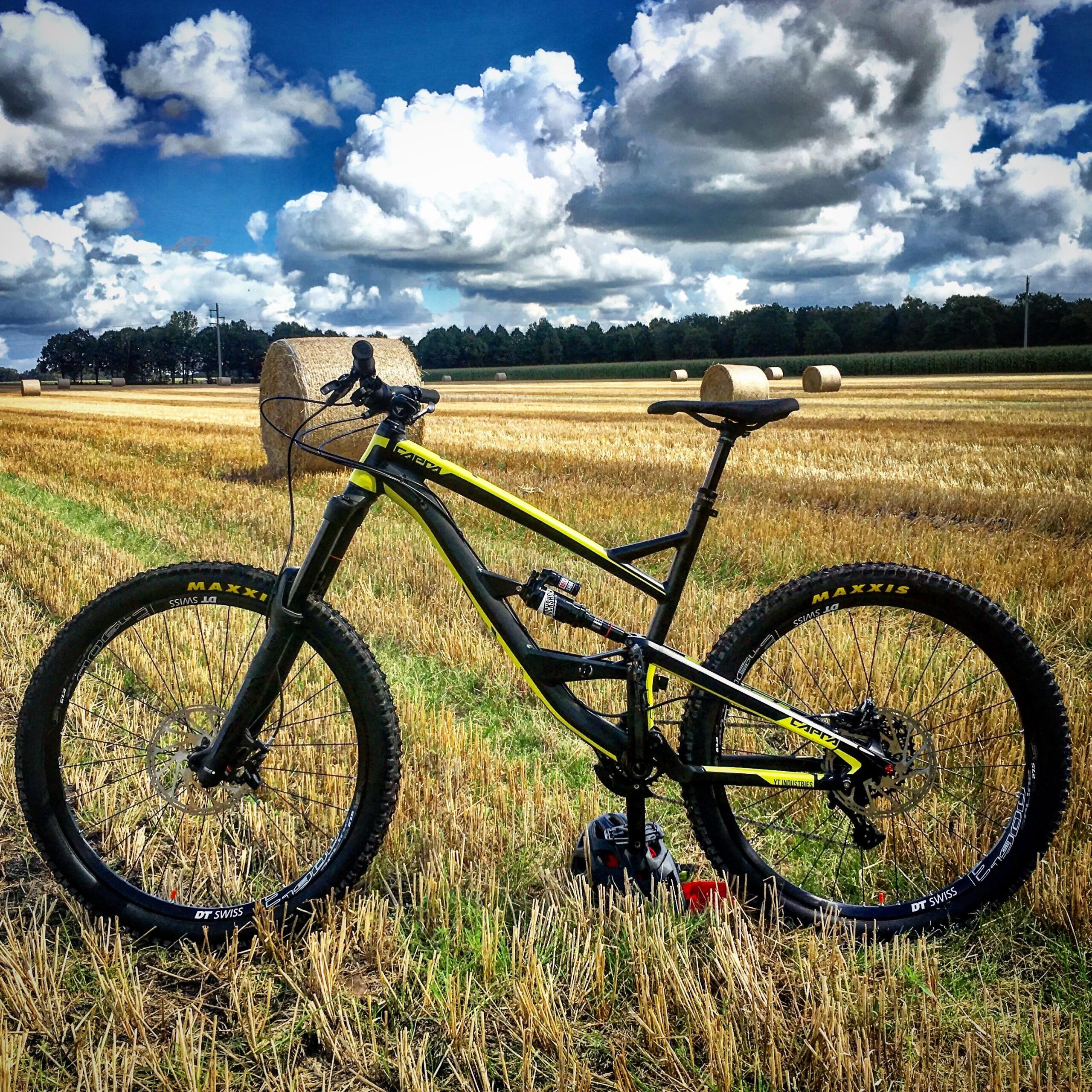 YT Industries Capra AL: A black and yellow mountain bike is positioned on a field of harvested straw, with round hay bales scattered in the background. The sky is partly cloudy, showcasing a mix of blue and white clouds. The bike features thick, knobby tires designed for off-road terrain.