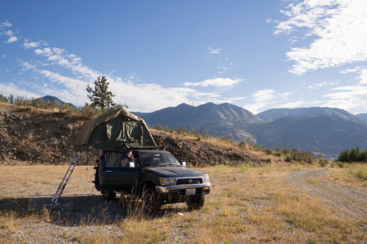 A green pickup truck with a rooftop tent is parked in an open field surrounded by mountains. A person is sitting inside the truck, with a ladder leaning against the vehicle leading up to the tent. The sky is clear with a few clouds, and the landscape features dry grasses and sparse trees.