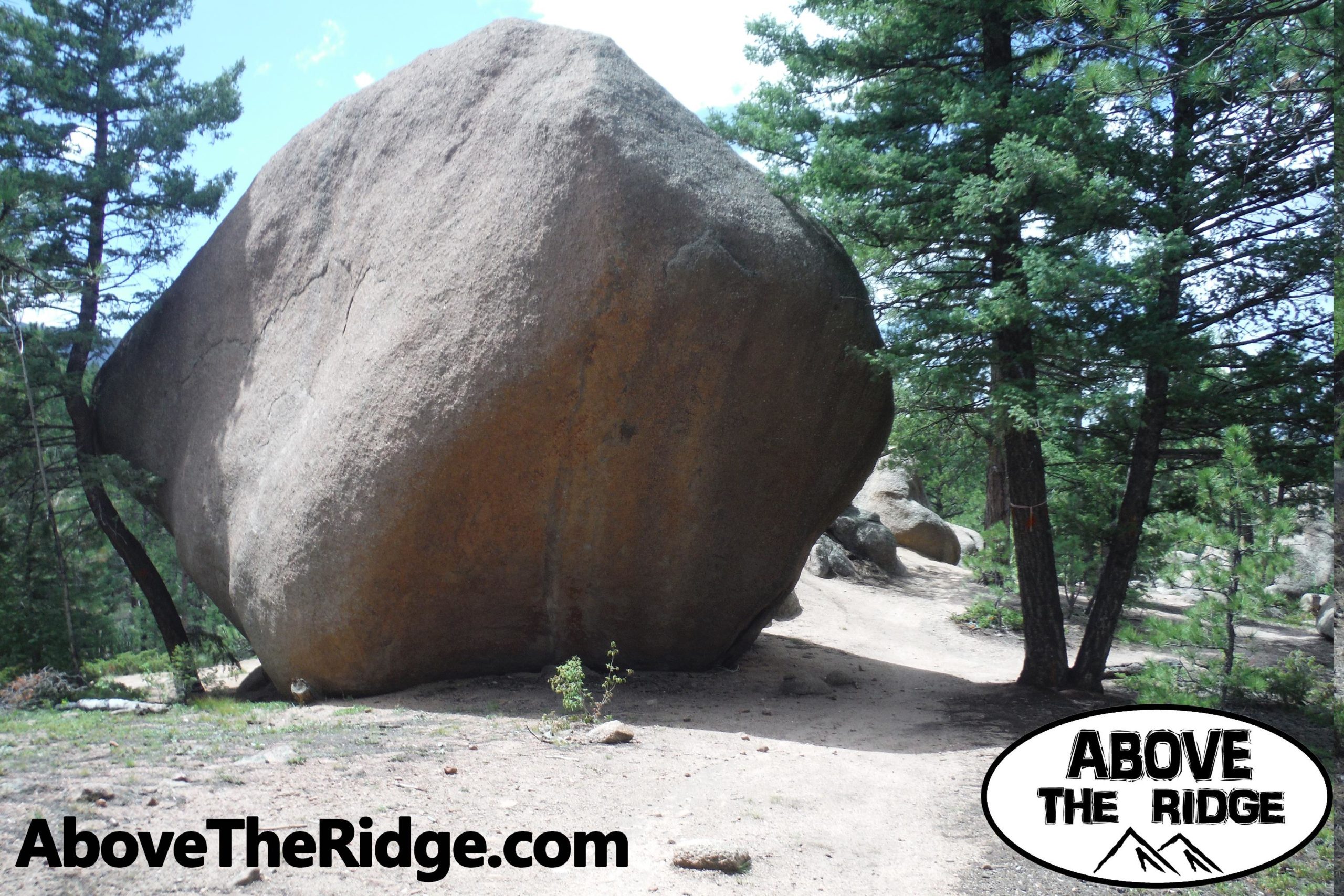 A large boulder stands prominently among green pine trees in a natural landscape. A dirt path curves around the boulder, inviting exploration. The image includes a logo in the bottom right corner that reads "ABOVE THE RIDGE," suggesting the theme of outdoor adventure. Little Scraggy mountain bike trail.