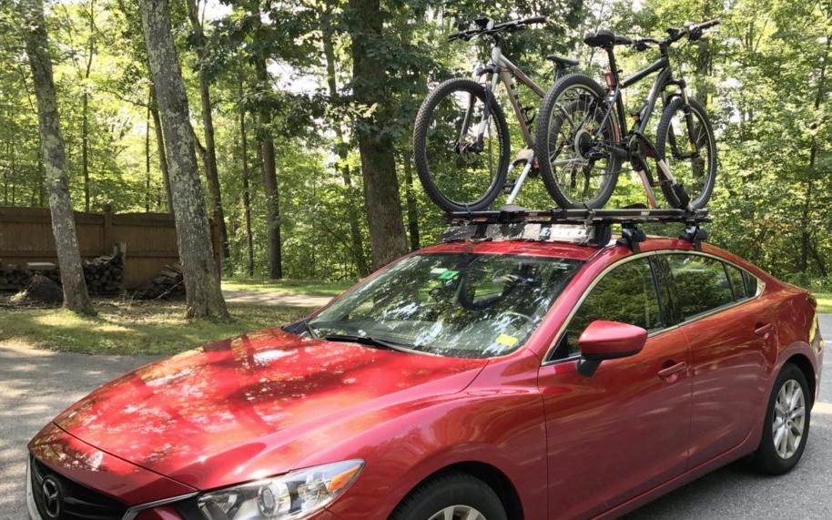Trek Marlin 5: A red car parked on a road surrounded by trees, with two mountain bikes secured on a roof rack. The setting indicates a recreational area, ideal for biking.