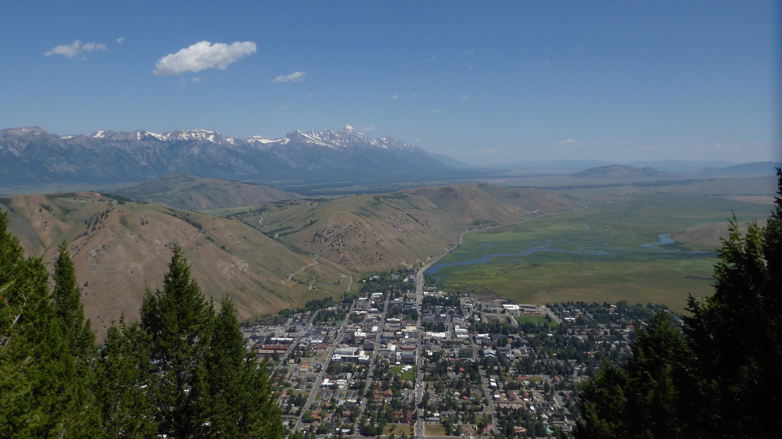 A panoramic view of a valley with a small town nestled among rolling hills, surrounded by mountains. The scene features green fields, a winding river, and a clear blue sky with a few clouds overhead. The foreground shows treetops, adding a natural frame to the landscape. Snow King Trail mountain bike trail.