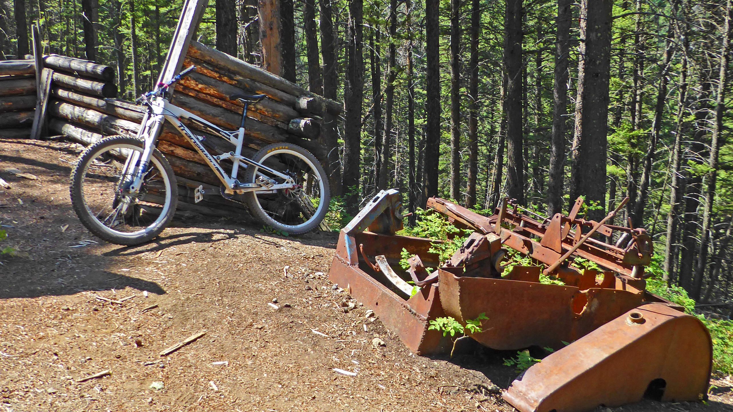 A mountain bike parked next to an old, rusted piece of machinery in a wooded area, with logs stacked in the background and trees surrounding the scene. Leverich Trail mountain bike trail.