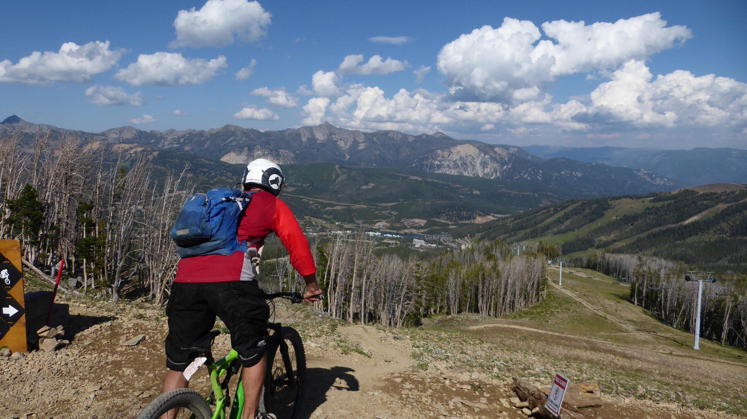 A mountain biker in a red shirt and a helmet is poised on a trail overlooking a valley and distant mountains under a blue sky with clouds. In the foreground, a sign marks the trail, while the landscape features sparse trees and a ski lift in the background. Soul Hole mountain bike trail.
