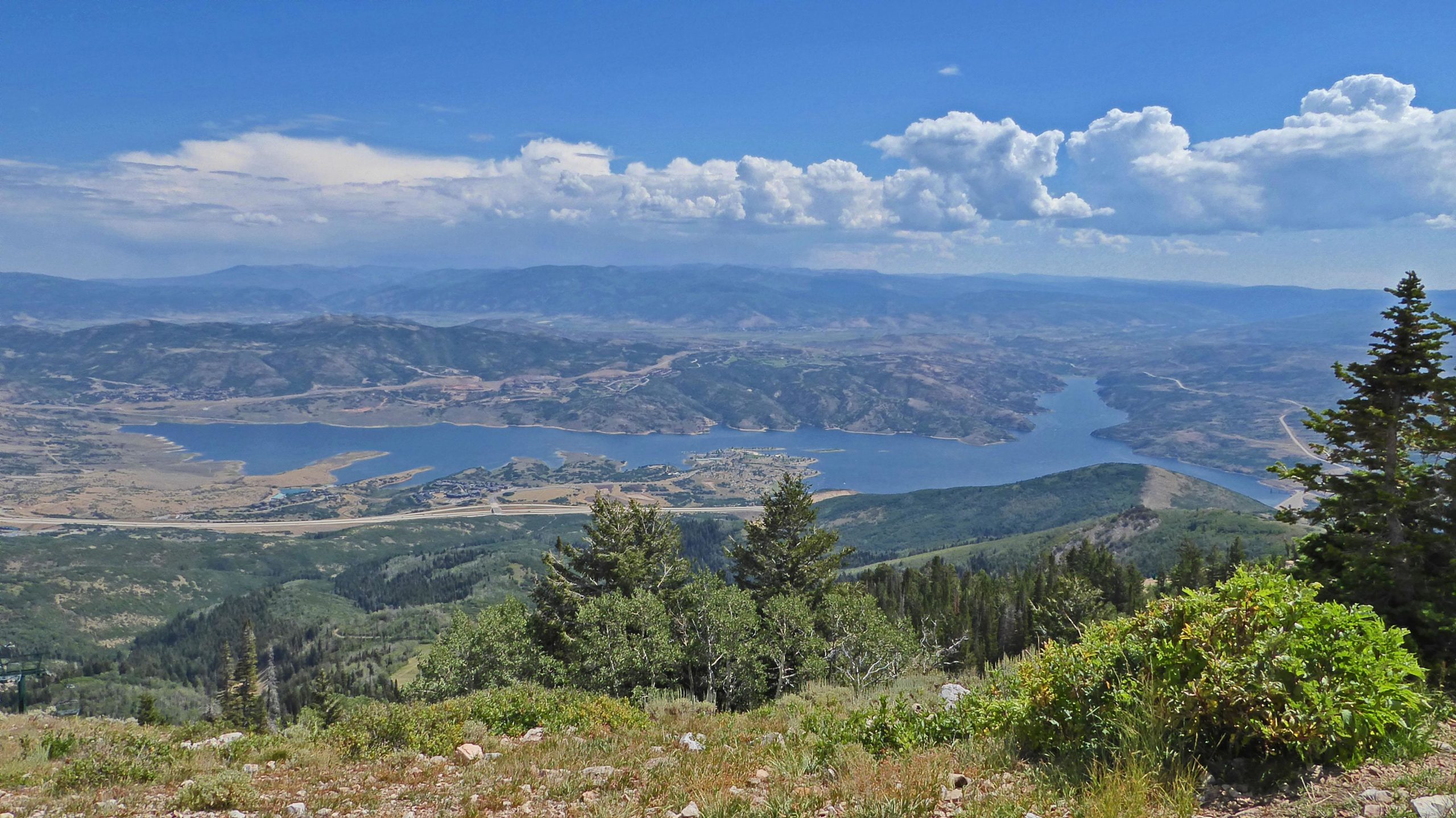 A scenic view from a mountain overlooking a large lake surrounded by green hills, with distant mountains under a blue sky dotted with clouds. Deer Valley Resort Bike Park mountain bike trail.