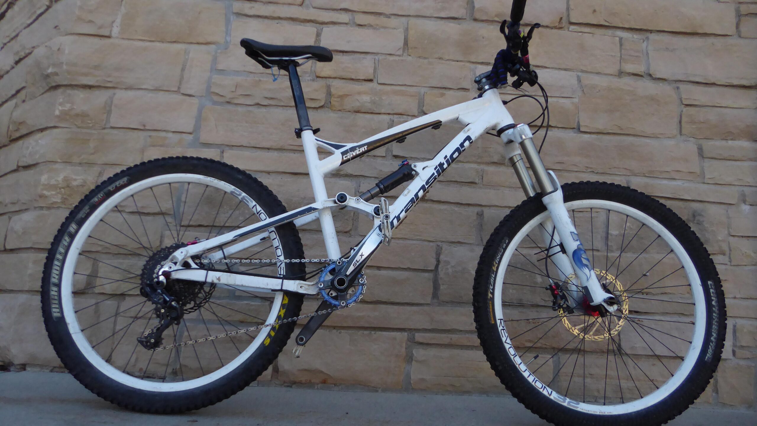 Transition Covert: A white mountain bike with black and blue accents, positioned against a stone wall. The bike features large, knobby tires designed for off-road terrain, a prominent suspension fork, and a modern frame design. The chain and gears are visible, showcasing its functionality for rugged trails.
