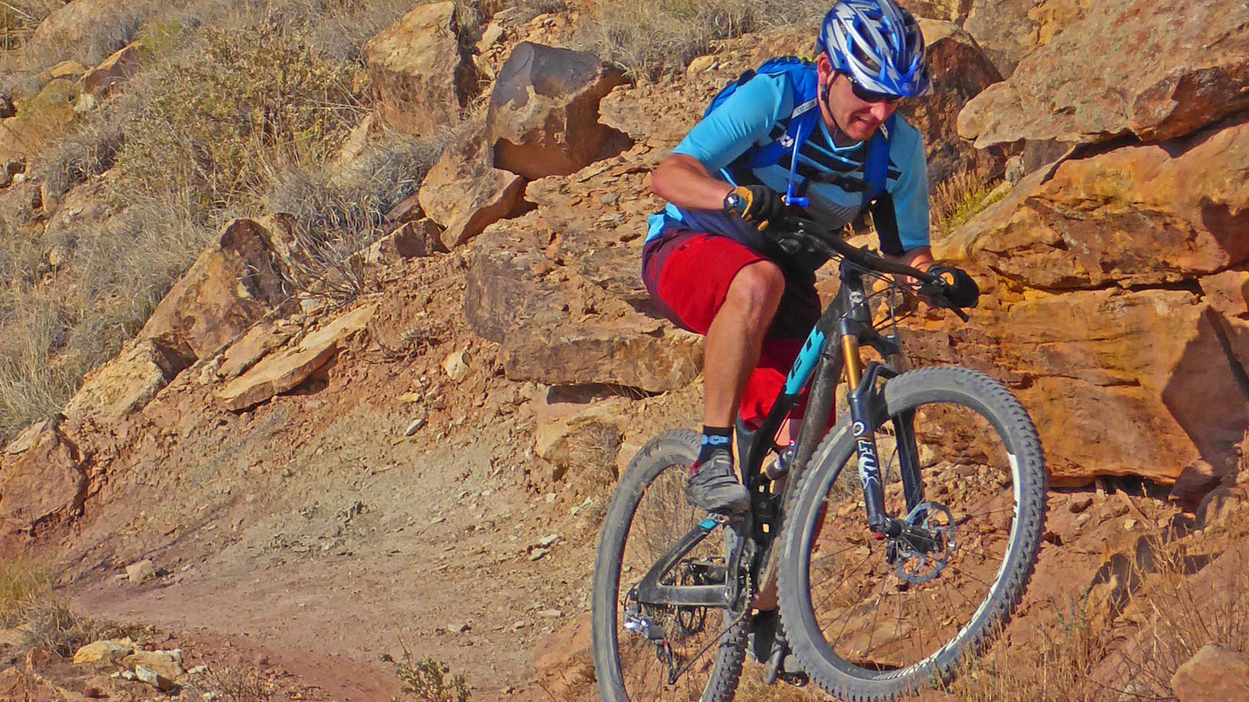 A mountain biker performing a jump on a rocky terrain, wearing a blue helmet, a blue shirt, and red shorts. The background features a landscape of rocks and sparse vegetation. Lunch Loops mountain bike trail.