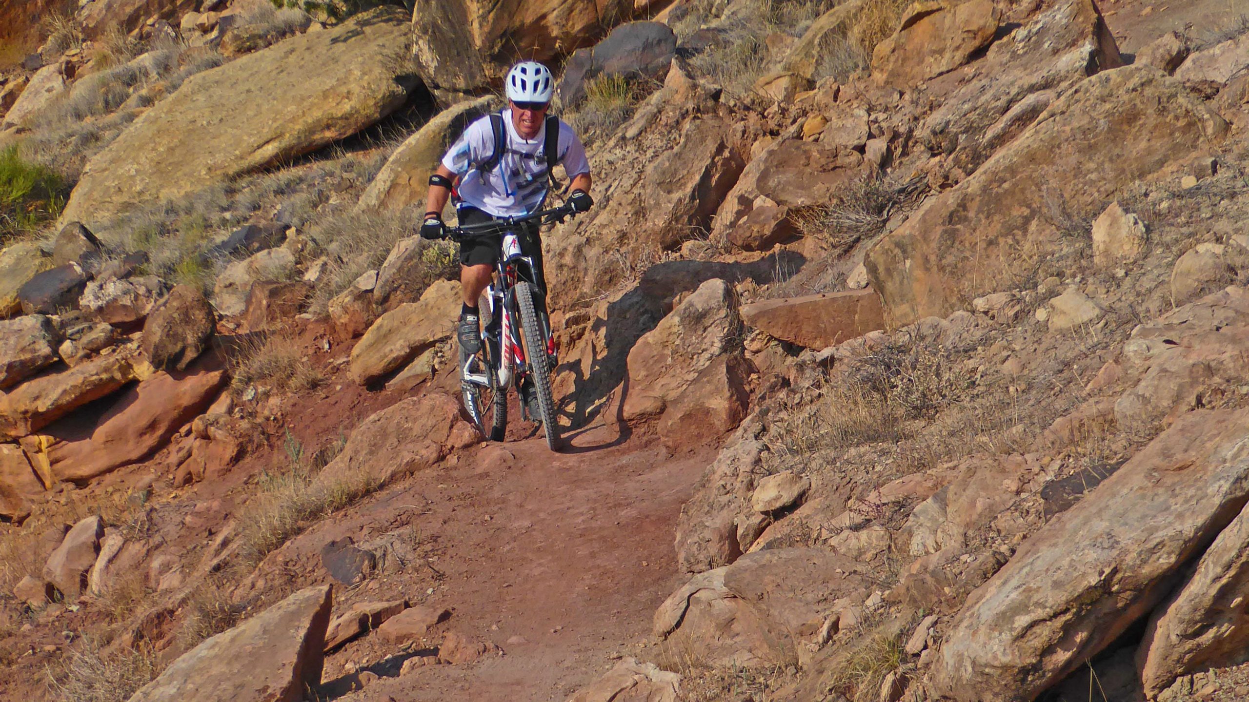 A mountain biker navigating a rocky trail, wearing a helmet and protective gear, with a rugged landscape of rocks and dry grass in the background. Lunch Loops mountain bike trail.