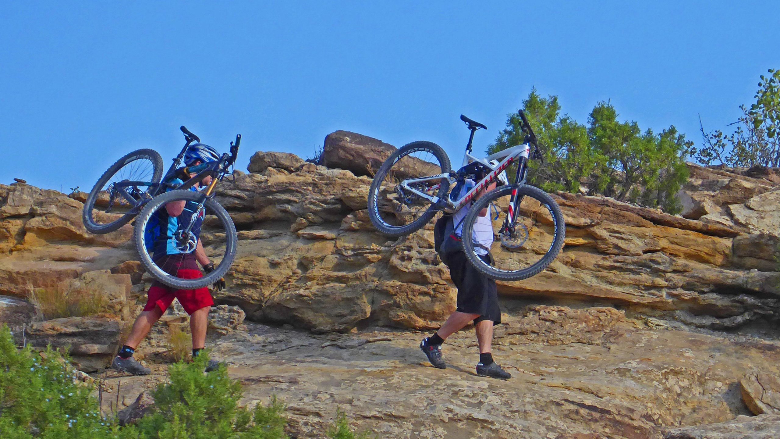 Two mountain bikers are walking uphill along a rocky terrain, each carrying a bicycle over their shoulders. The background features rugged stones and sparse vegetation under a clear blue sky. Lunch Loops mountain bike trail.