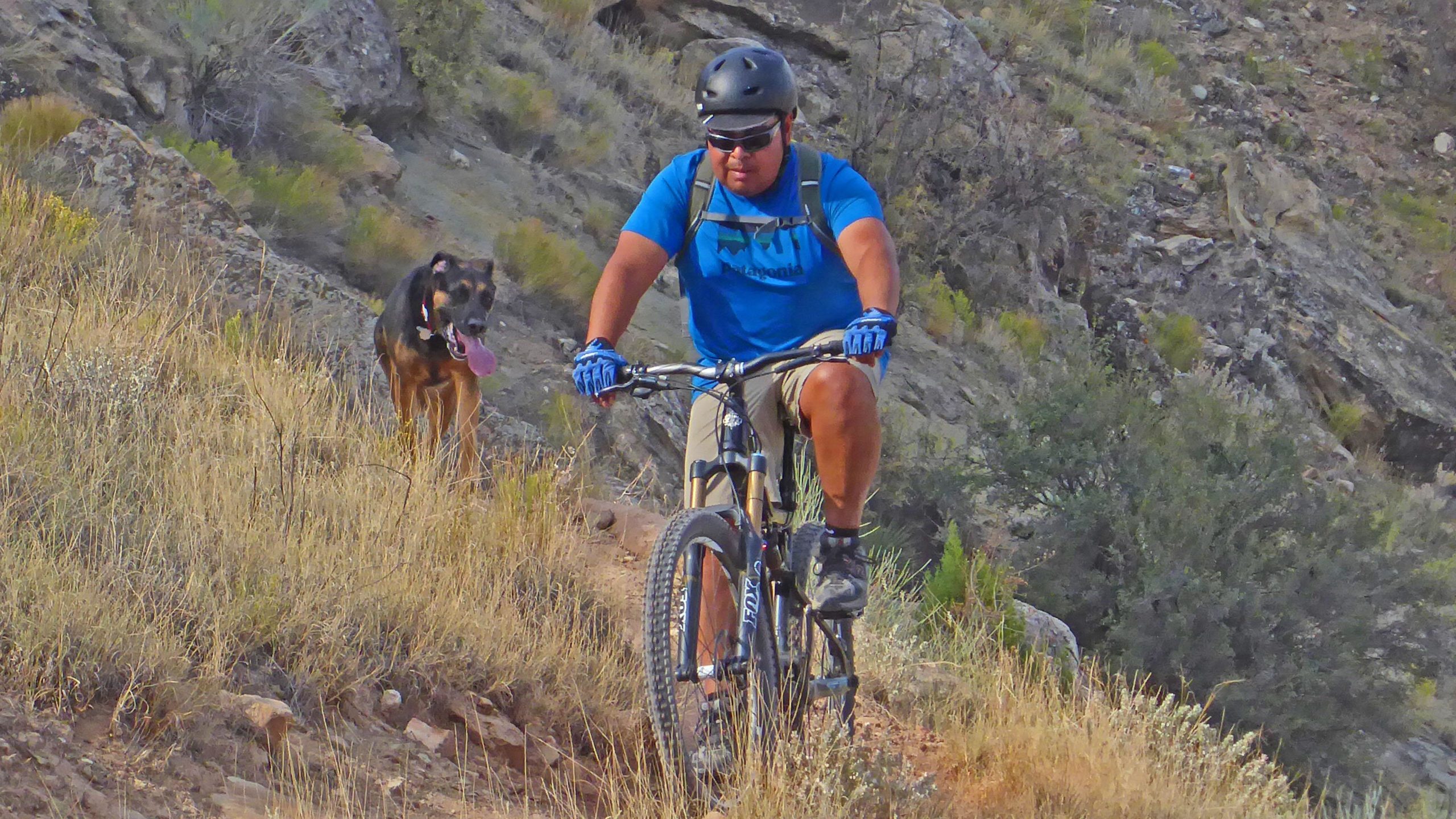 A person wearing a blue shirt, helmet, and gloves rides a mountain bike on a rocky trail alongside a playful dog with its tongue out. The scene features dry grass and vegetation, with rugged terrain and rocks in the background. Lunch Loops mountain bike trail.