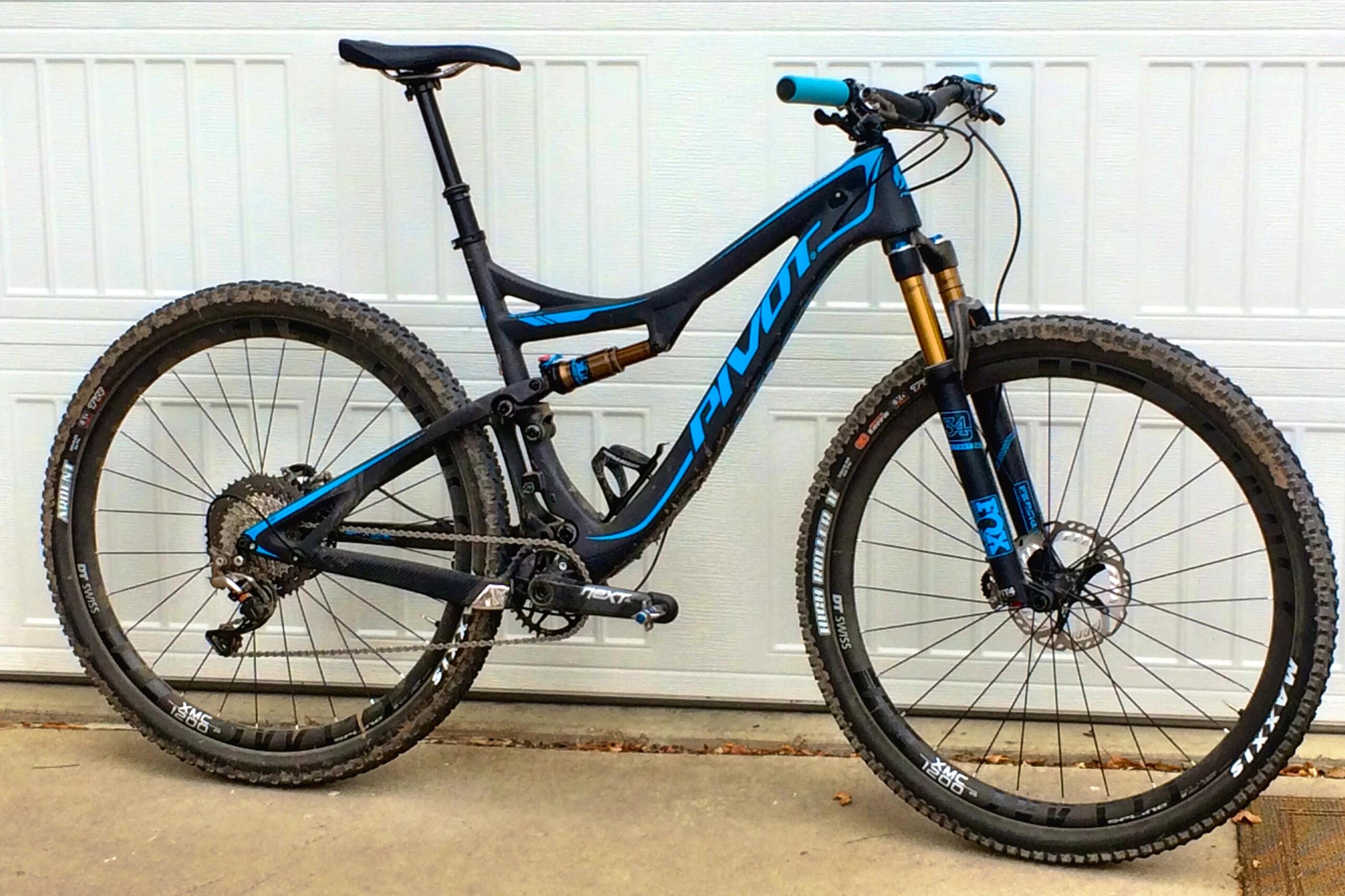 Pivot Mach 429SL Carbon: A black and blue mountain bike leaning against a garage door, showcasing its sleek frame, suspension components, and knobby tires. The bike features a dropper post, disc brakes, and a high-performance drivetrain suitable for off-road trails.