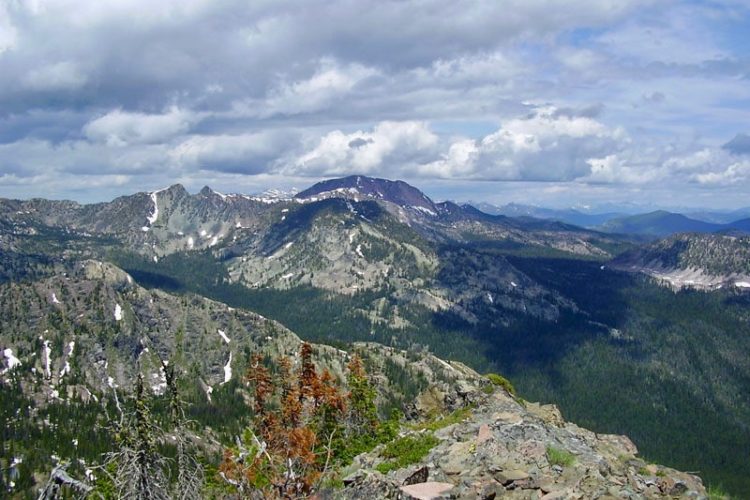 A panoramic view of rugged mountains under a partly cloudy sky, featuring a mix of rocky terrain and dense evergreen forests. Snow patches are visible on some mountain slopes, and the landscape stretches into the distance, showcasing multiple peaks and valleys.