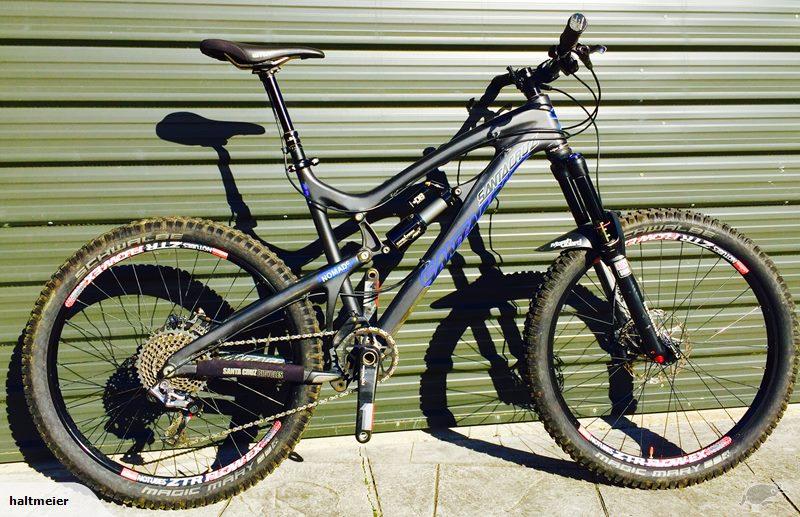 Santa Cruz Nomad Carbon: A black and blue mountain bike with thick tires, positioned against a green metal wall. The bike features a full suspension frame, front fork suspension, and various gears, showcasing its design for off-road cycling. The tires are slightly muddy, indicating recent use.