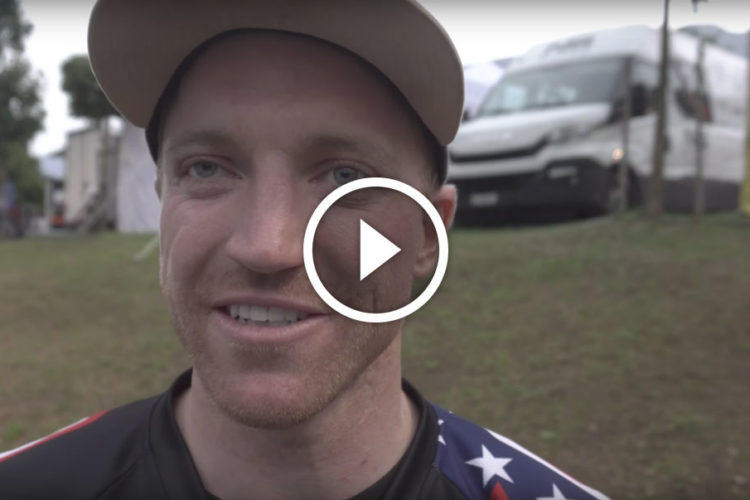 A close-up of a smiling man wearing a black shirt with red and blue accents, standing outdoors with green grass and vehicles in the background. A play button overlay indicates a video is available.