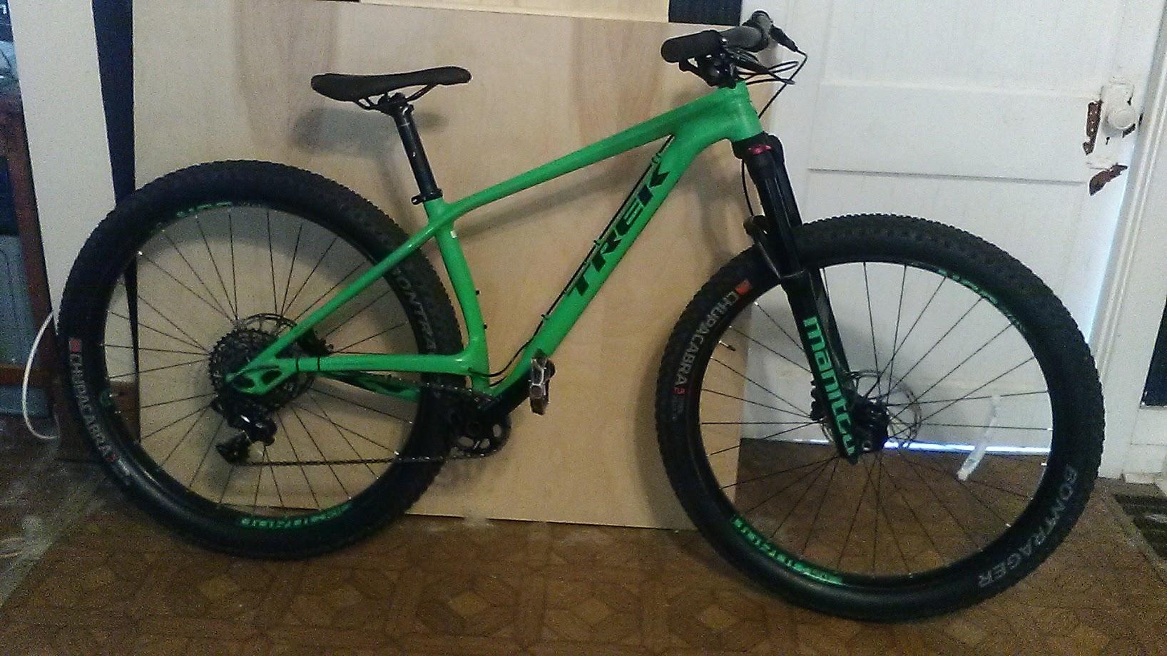 Trek Stache: A bright green mountain bike with large tires and a black front fork is leaning against a light-colored wooden panel. The bike features noticeable branding and has a modern design suitable for off-road riding. The background includes a door and some flooring details.