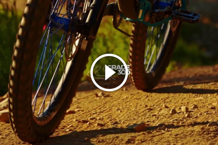 Close-up view of a mountain bike tire and wheel on a dirt trail, with sunlight illuminating the scene. The focus is on the textured tire, showing detail in the tread and surrounding gravel, suggesting an active outdoor environment. A play button overlay indicates that this is a video clip related to mountain biking.