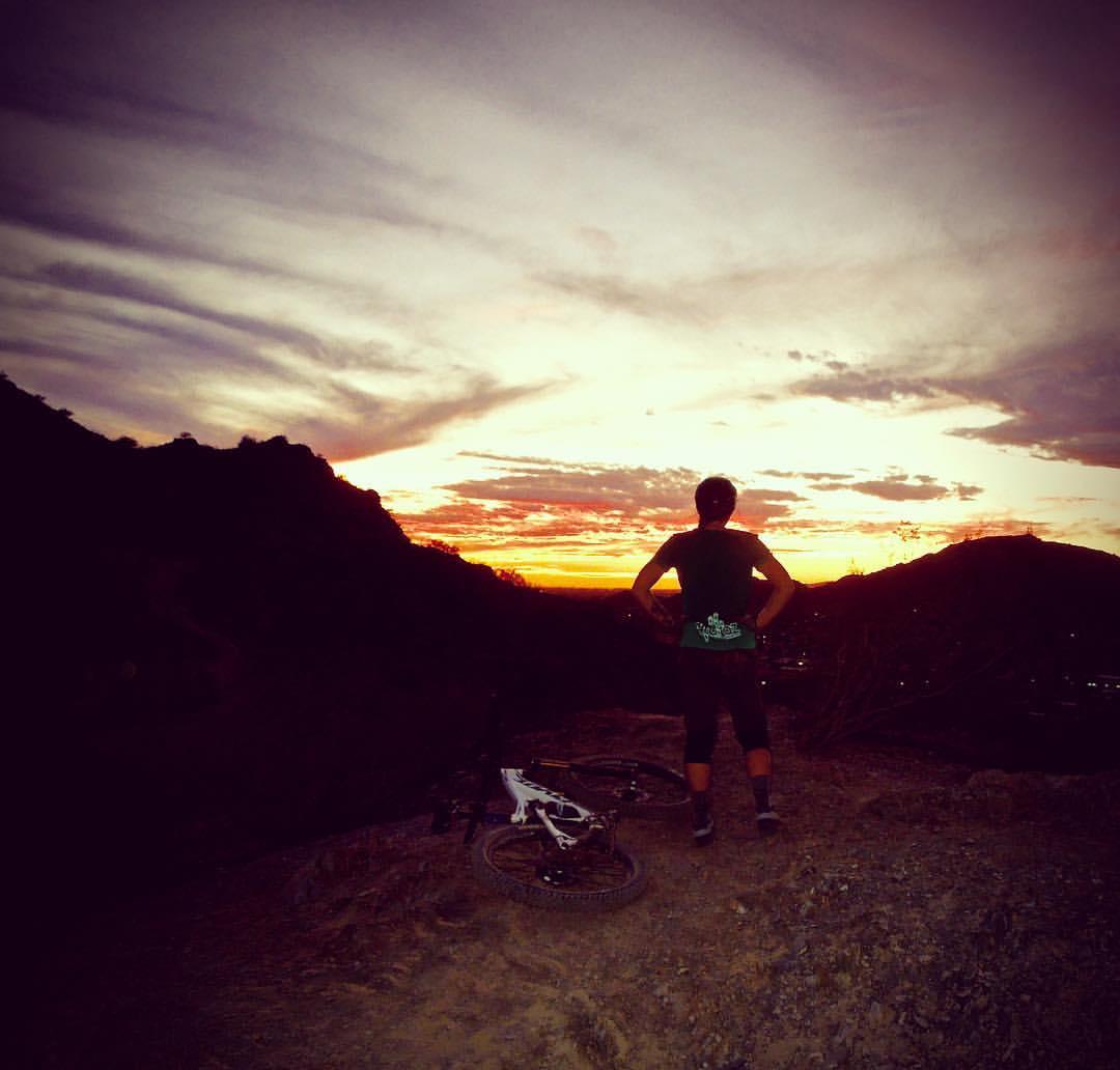 A person stands on a rocky ledge, silhouetted against a vibrant sunset with hues of orange, purple, and pink. A mountain bike rests on the ground nearby, and the landscape features rolling hills and a distant view of city lights below. The individual gazes thoughtfully at the horizon, creating a serene and contemplative atmosphere. Trail #100 mountain bike trail.